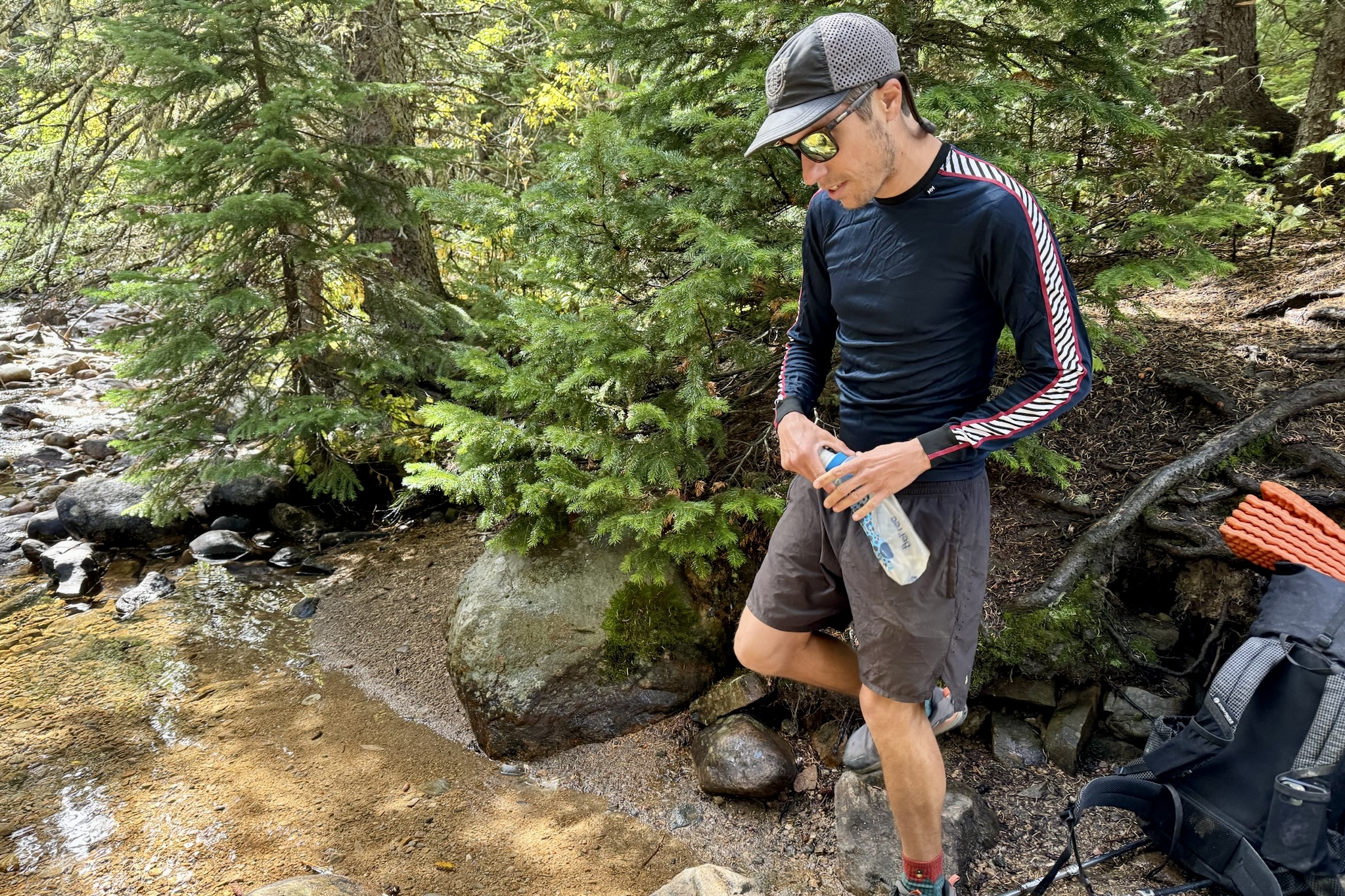 A hiker holds a water filter while wearing the Helly Hansen Lifa Stripe base layer. Trees, a creek, and a backpack are in the background.