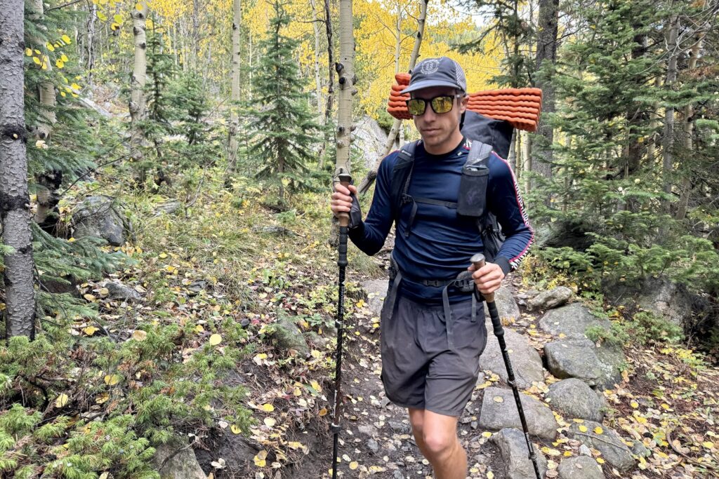 A hiker hikes towards the camera with a pack and trekking poles while wearing the Helly Hansen Lifa Stripe base layer. Trees are in the background.