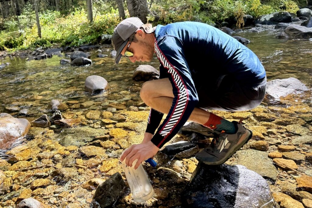 A hiker crouches by a creek to fill a water bottle while wearing the Helly Hansen Lifa Stripe base layer. Rocks and trees are in the background.