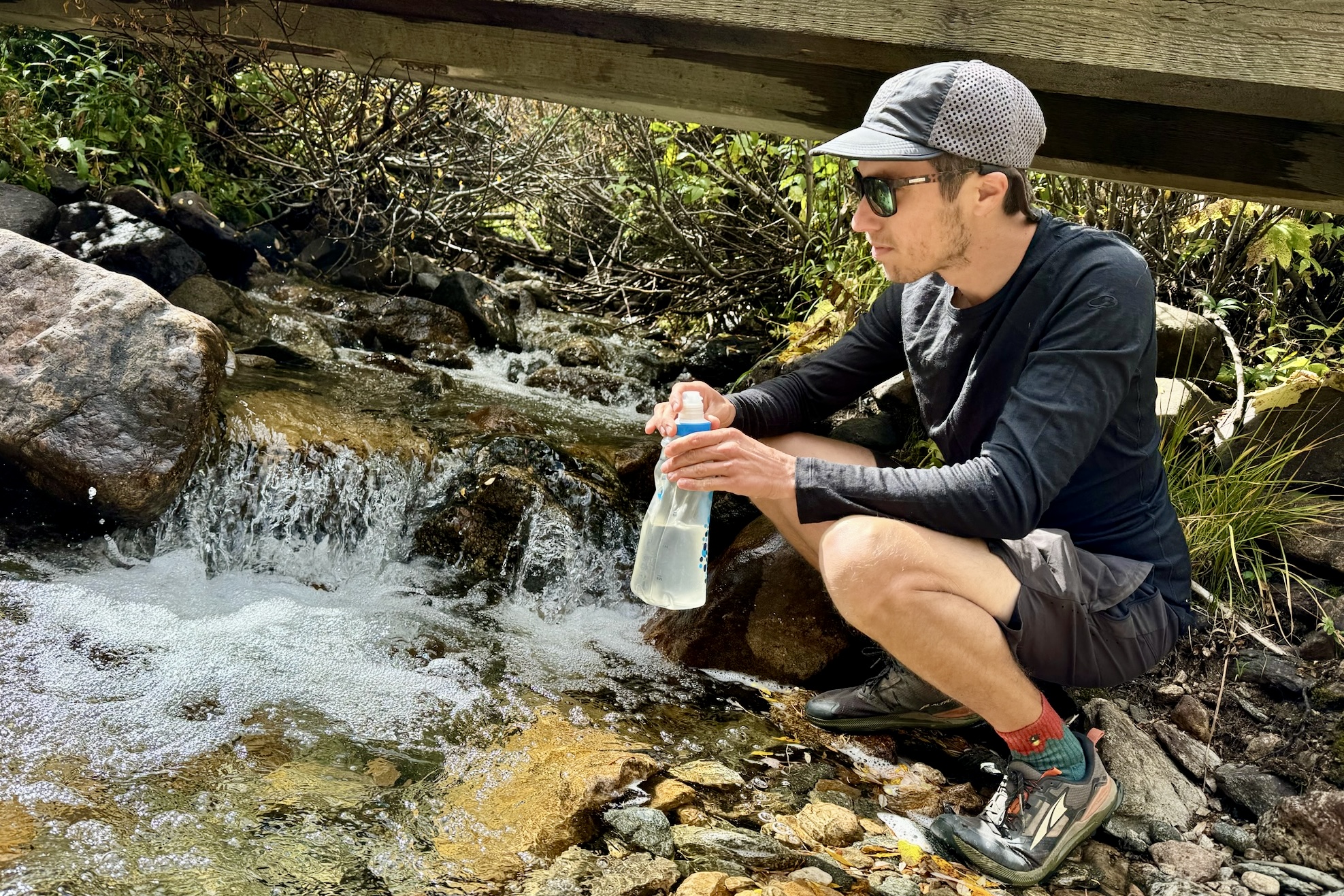 A hiker fills a water bladder by a creek while crouching and wearing the Icebreaker Oasis 200 base layer.