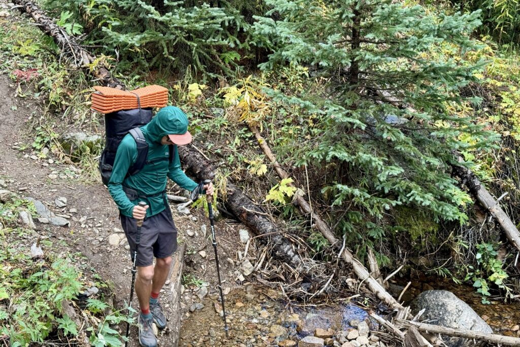 A hiker descending a steep trail while carrying a pack and wearing the Outdoor Research Echo Hoodie. Trees are in the background.