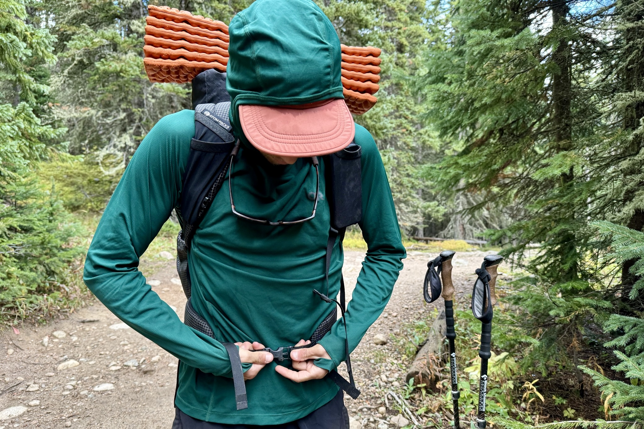 A hiker buckles the waist belt of his backpack while wearing the Outdoor Research Echo Hoodie. Trees, trekking poles, and a trail are in the background.