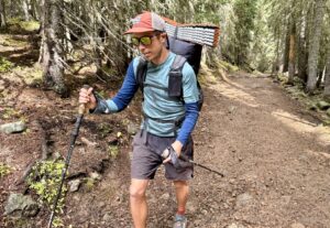 A hiker hiking through the woods with a pack and trekking poles while wearing the Ortovox Rock'n'Wool base layer. Trees and a trail are in the background.