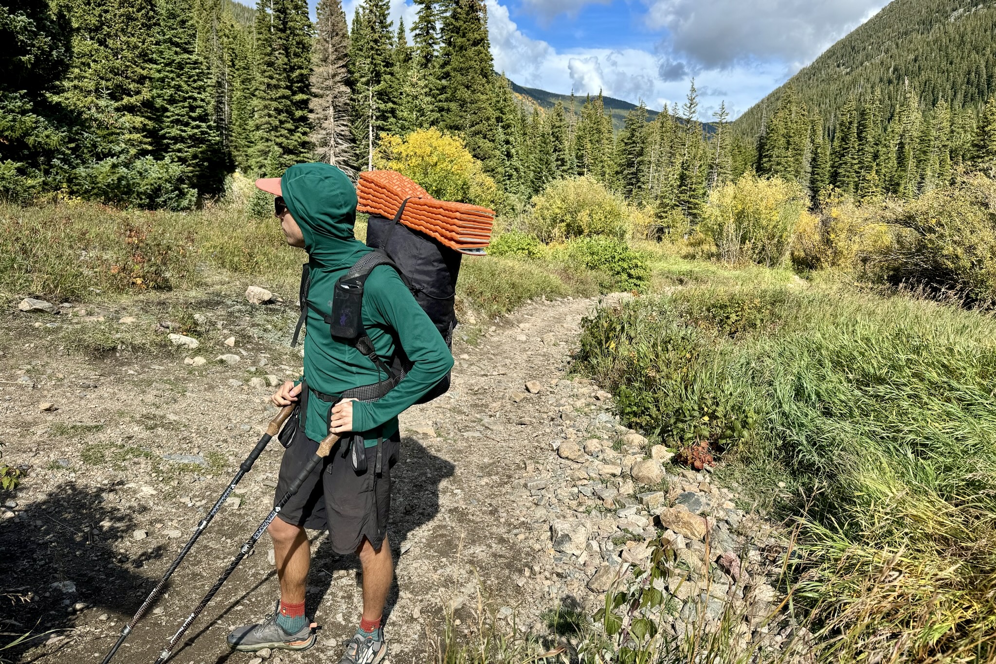 A hiker standing in the middle of a trail with trees and mountains in the background. He is leaning on his trekking poles while wearing a pack and the Outdoor Research Echo Hoodie.