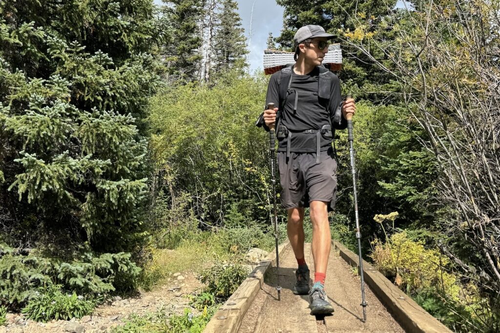 A hiker crosses a bridge with trekking poles, a pack, and wearing the Icebreaker Oasis 200 base layer. Trees are in the background.