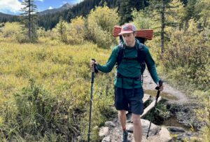 A hiker walks down a trail with mountains and trees in the background. He is using trekking poles and wearing a pack and the Outdoor Research Echo Hoodie