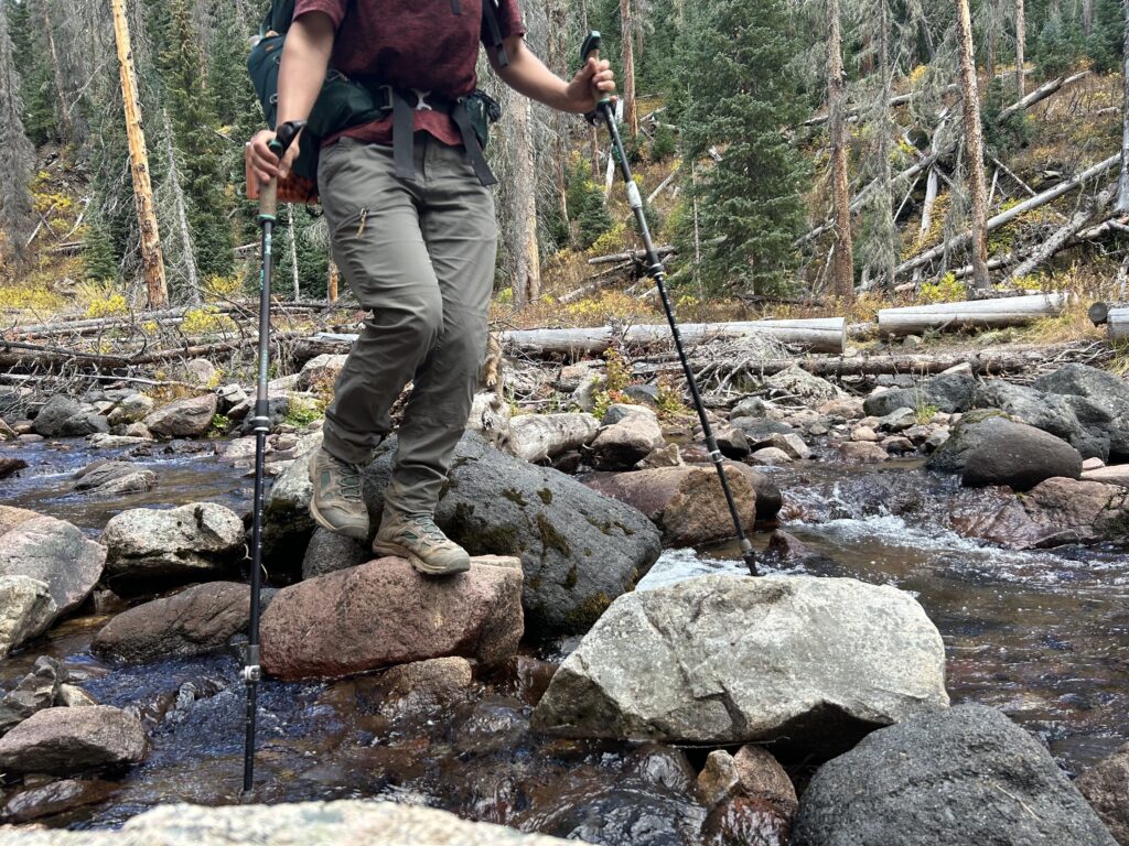 A hiker wearing the REI Trailmade Pants crosses a stream on some rocks.