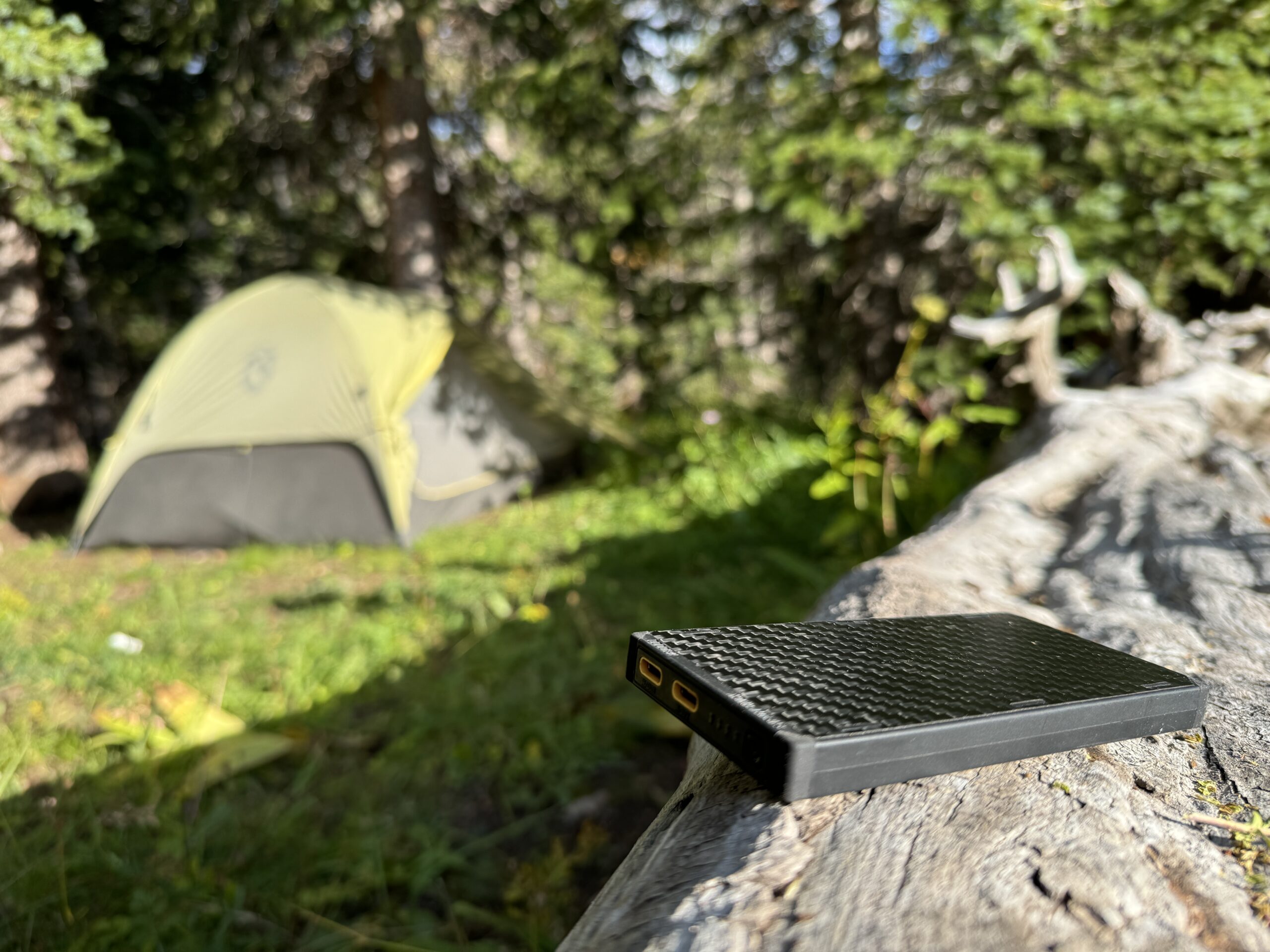 The Nitecore NB10000 power bank sitting on a log with a tent and trees in the background.