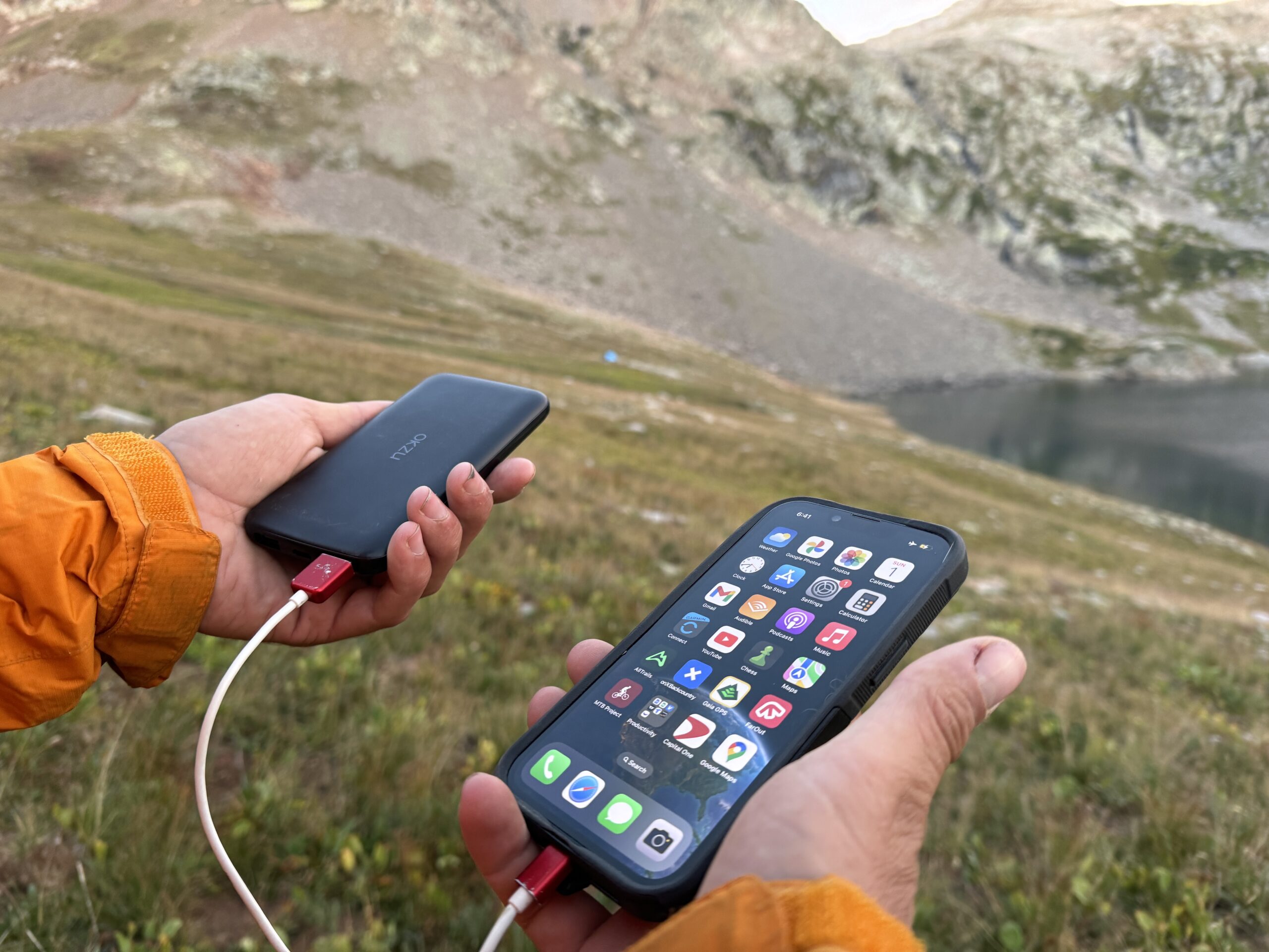 Close up of a phone and the OKZU power bank being held in a man's hands with a mountain lake in the background.