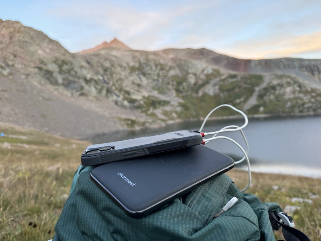 Close up of the Charmast power bank charging a phone that is sitting on a backpack with a lake and mountain in the background.