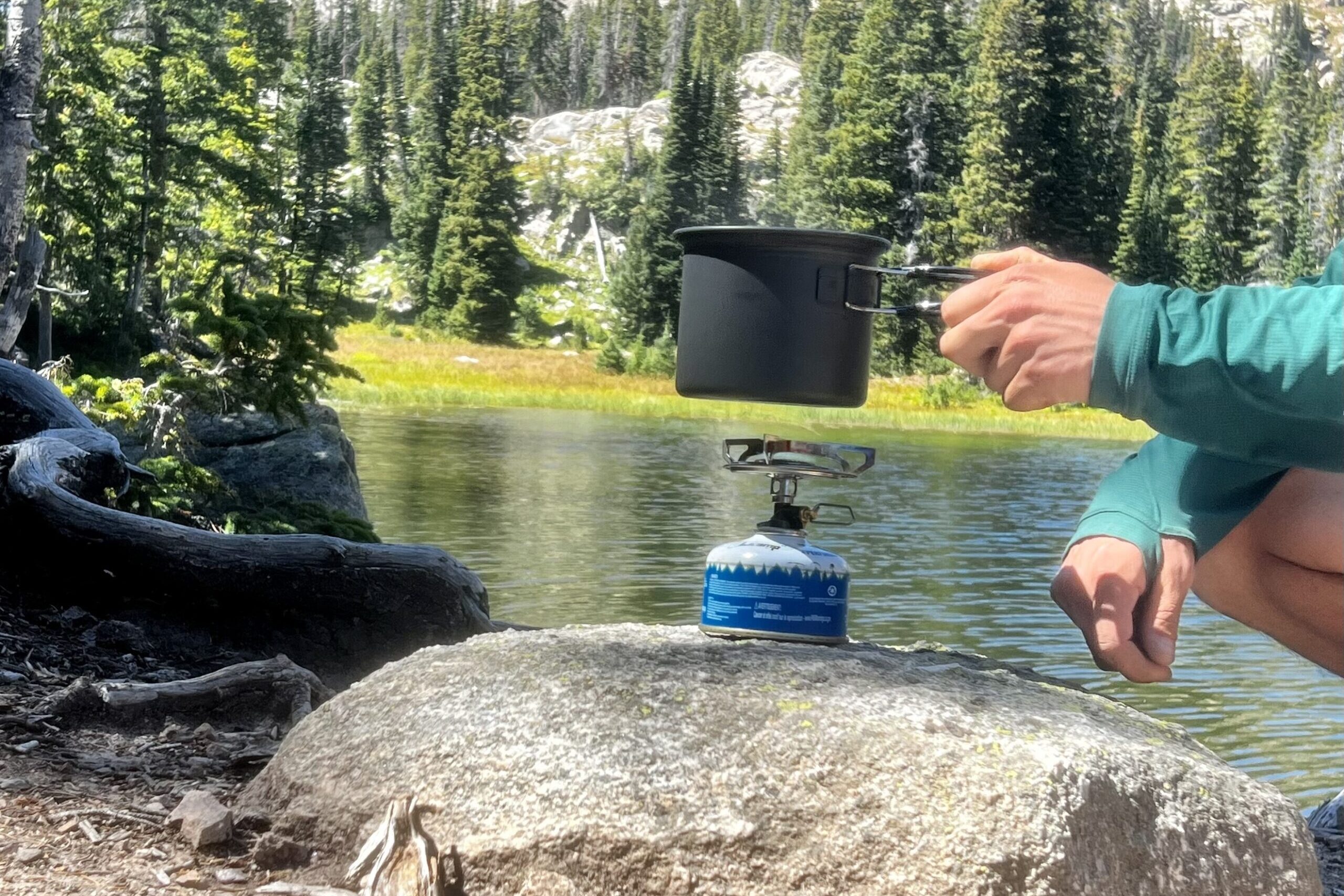 The hands of a hiker prepare to place a pot on the Primus Essential Trail while the flame is burning. The stove sits on a rock and the hiker his holding the pot above the stove. The background is a lake and trees.