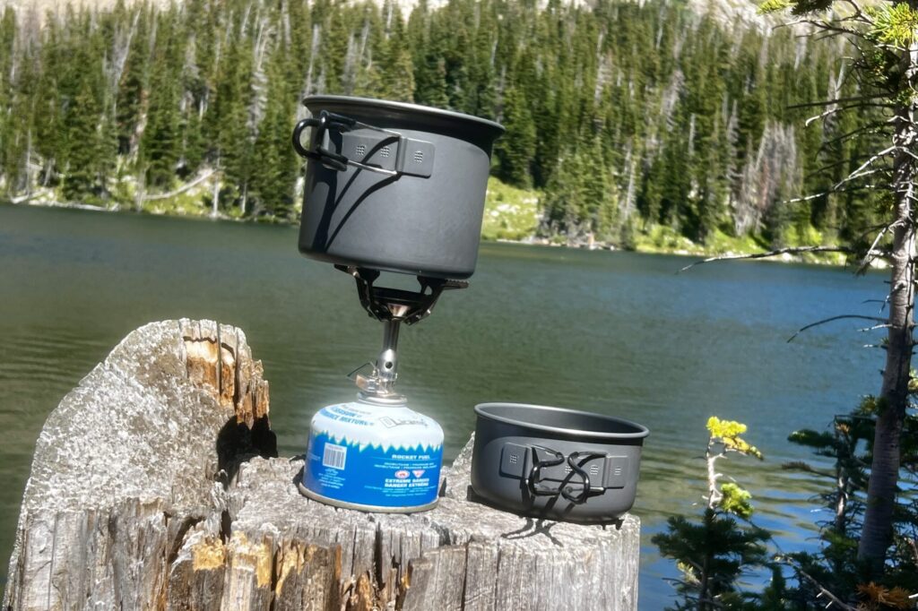 The SOTO Amicus cookset combo on a tree stump with a lake in the background. The main pot sits on the stove, ready to cook while the lid sits next to it on the stump.