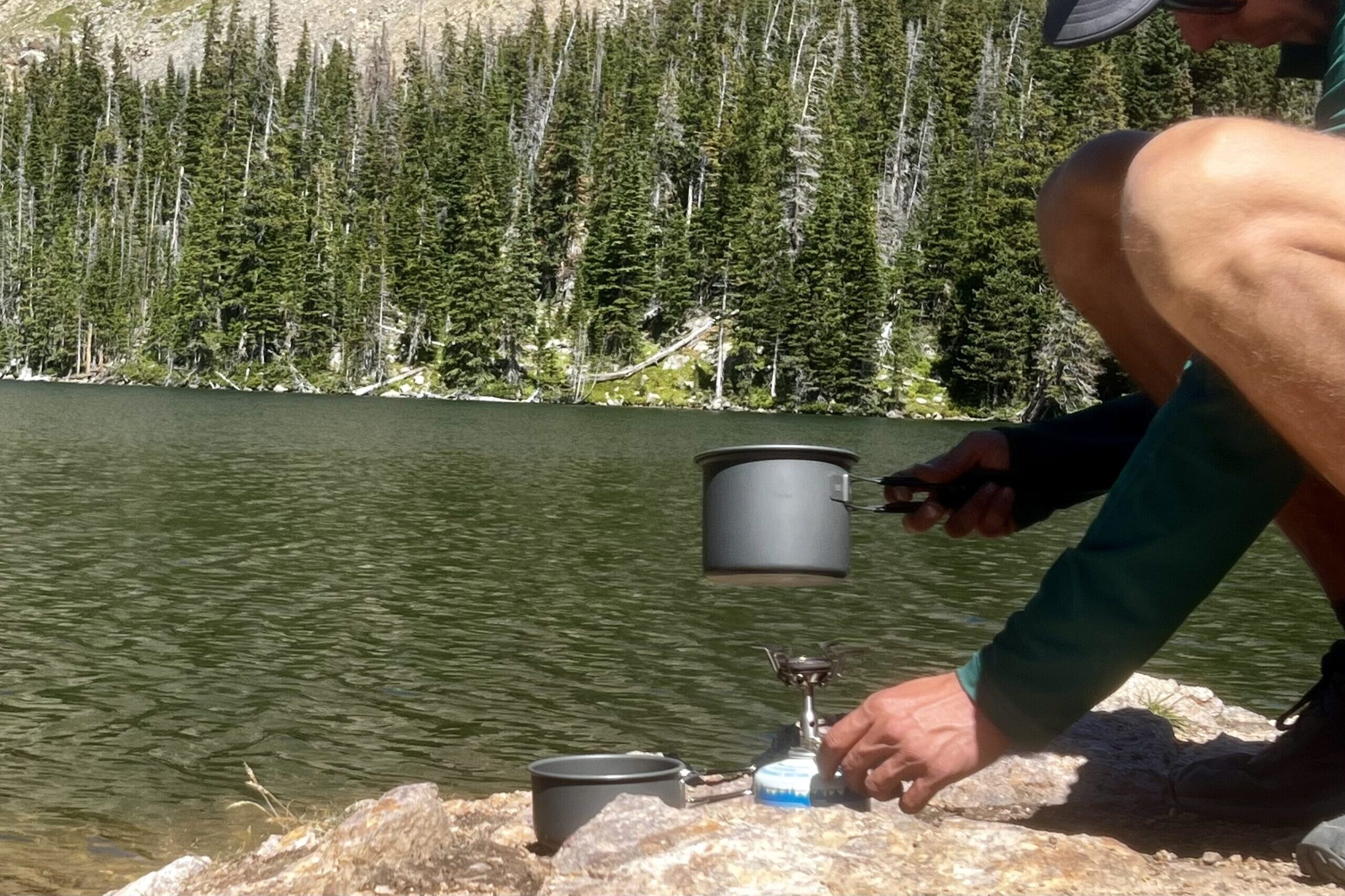 A hiker cooks with the SOTO Amicus cookset combo. He adjusts the flame of the stove while holding the pot a few inches above. The lid is nearby and the background features an alpine lake and trees.