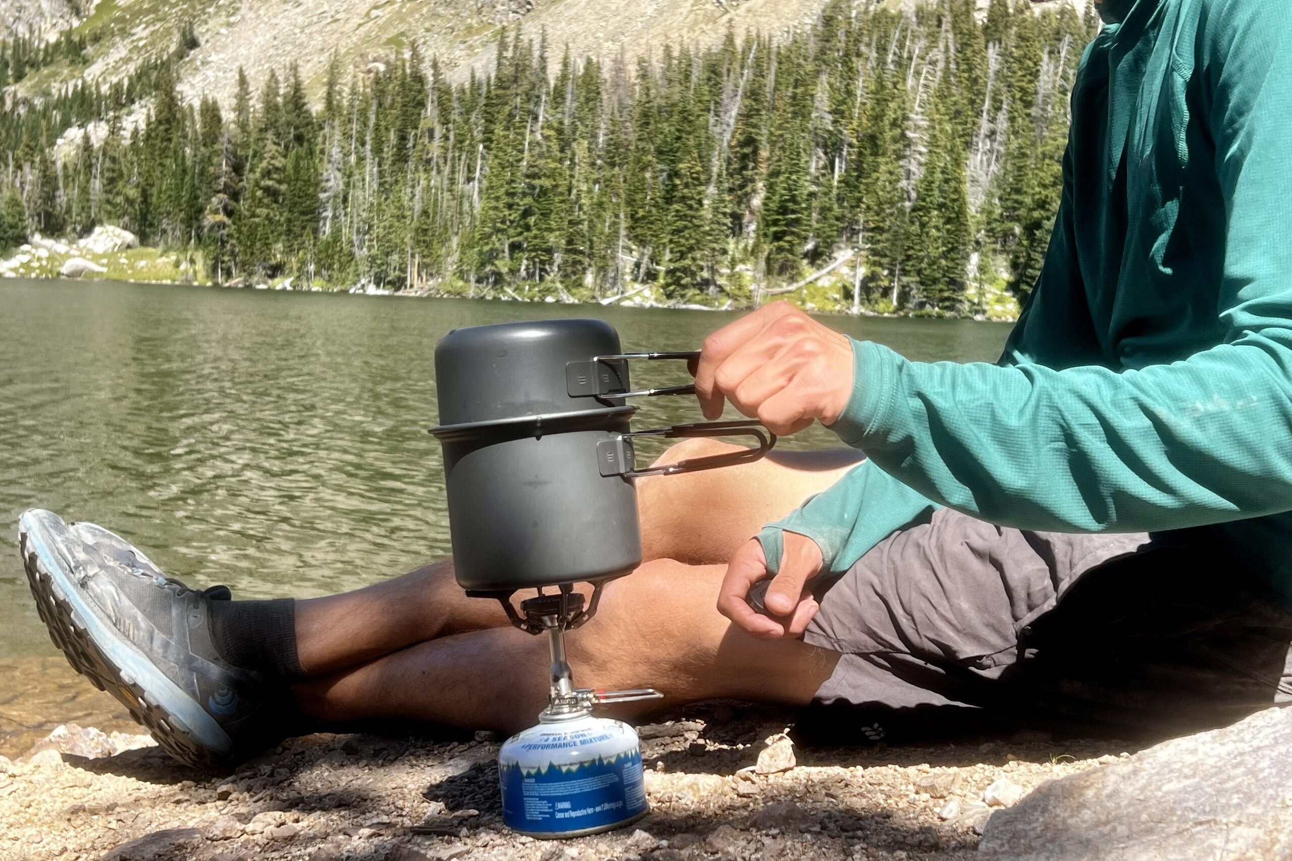 A hiker cooks with the SOTO Amicus Cookset Combo. The stove and fuel canister sits next to him, with the pot on the flame. He is placing the lid of the cookset combo on the main pot. There is a lake and trees in the background.