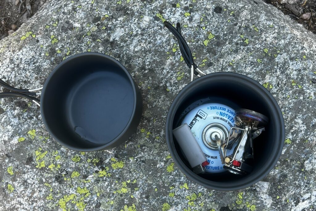 All the components of the SOTO Amicus Cookset Combo on a rock. The pan sits separate and the pot has a fuel canister, the stove without stuff sack, and mini bic light packed inside.