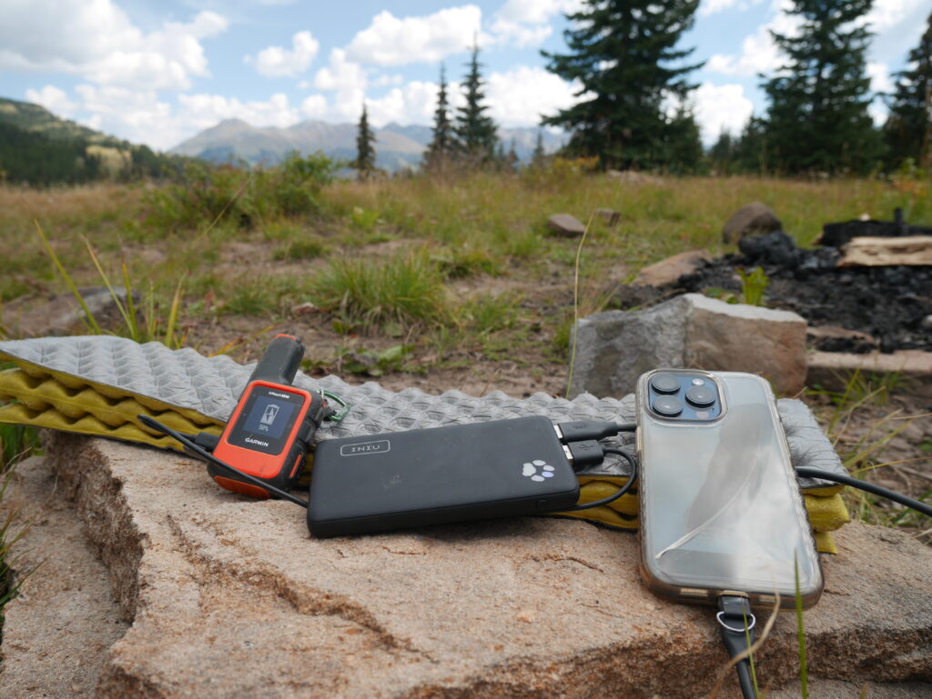 INIU Megapower 10000 charging a phone and the Garmin Inreach on a rock with trees and mountains in the background.