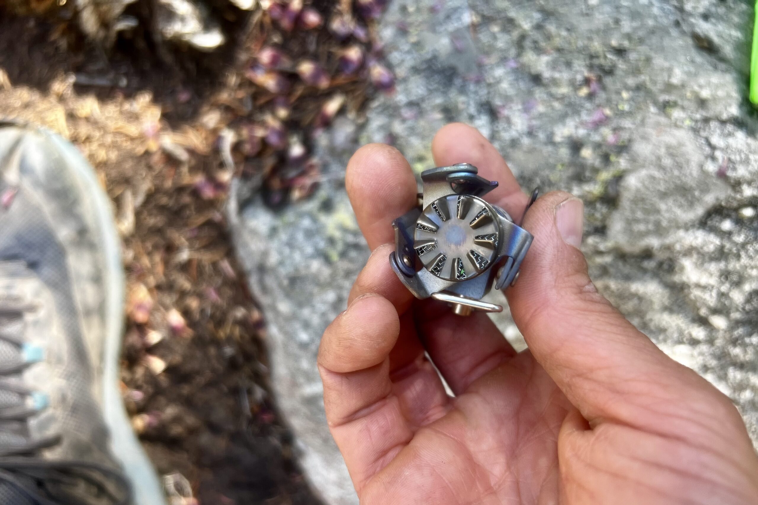 A hand holding the compacted BRS 3000T stove, showing a direct shot of its burner. Rocks and dirt in the background.