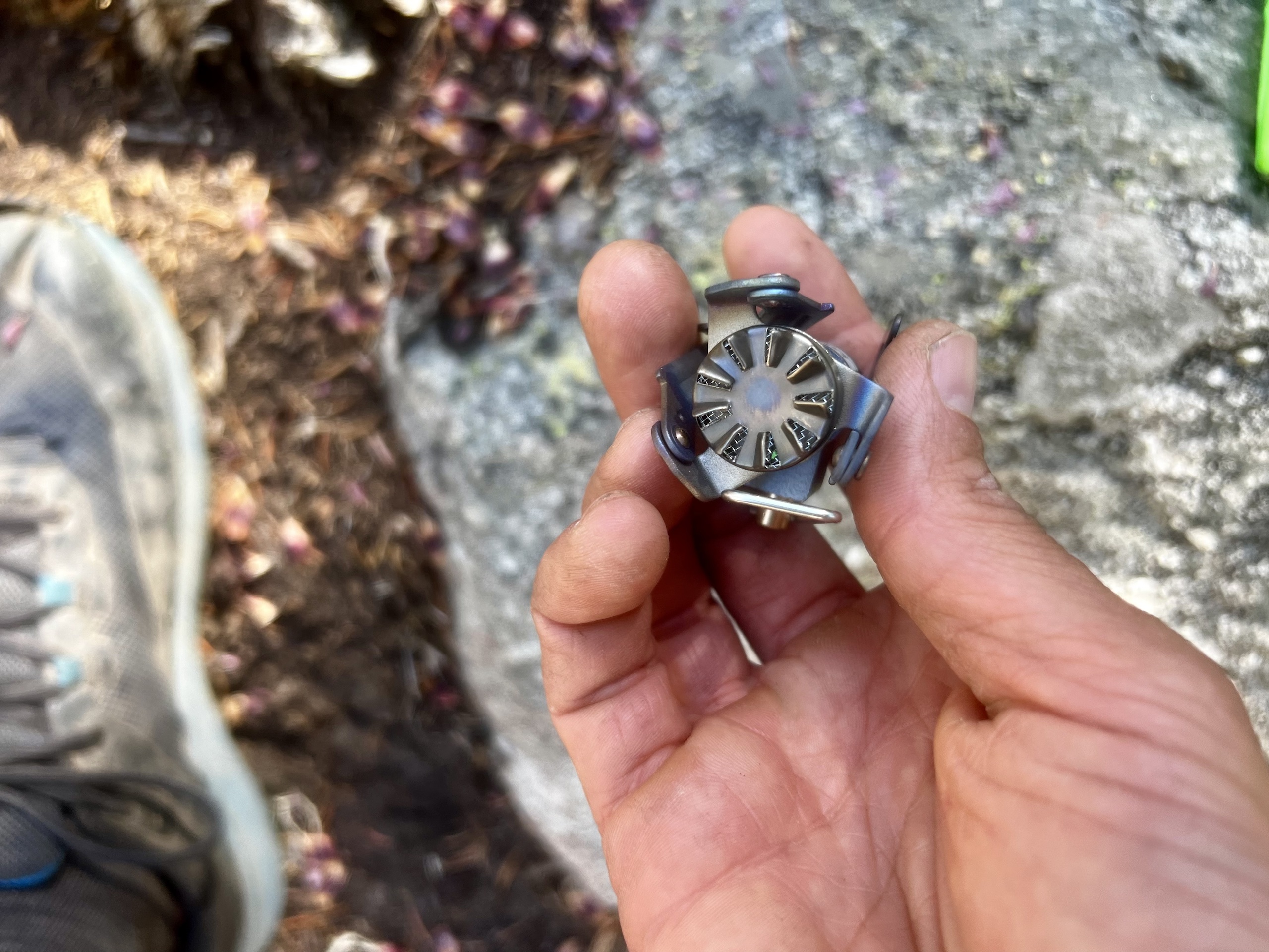 A hand holding the compacted BRS 3000T stove, showing a direct shot of its burner. Rocks and dirt in the background.