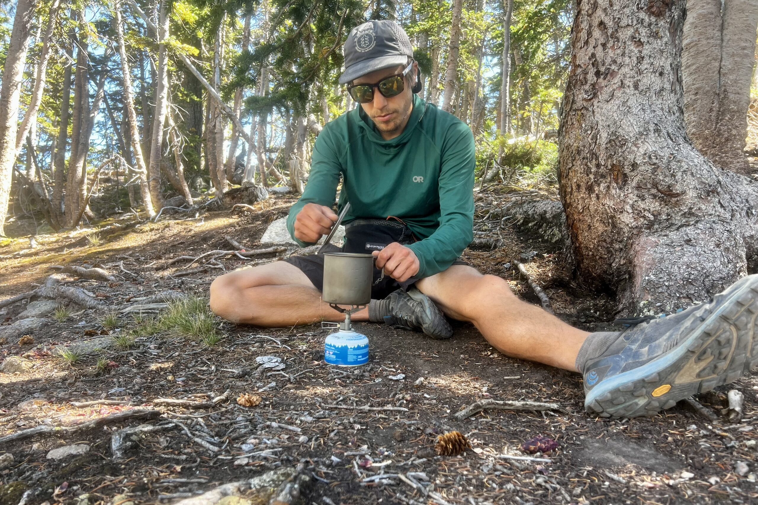 A hiker lifts the lid of his pot while cooking with the BRS 3000T in a wooded setting.