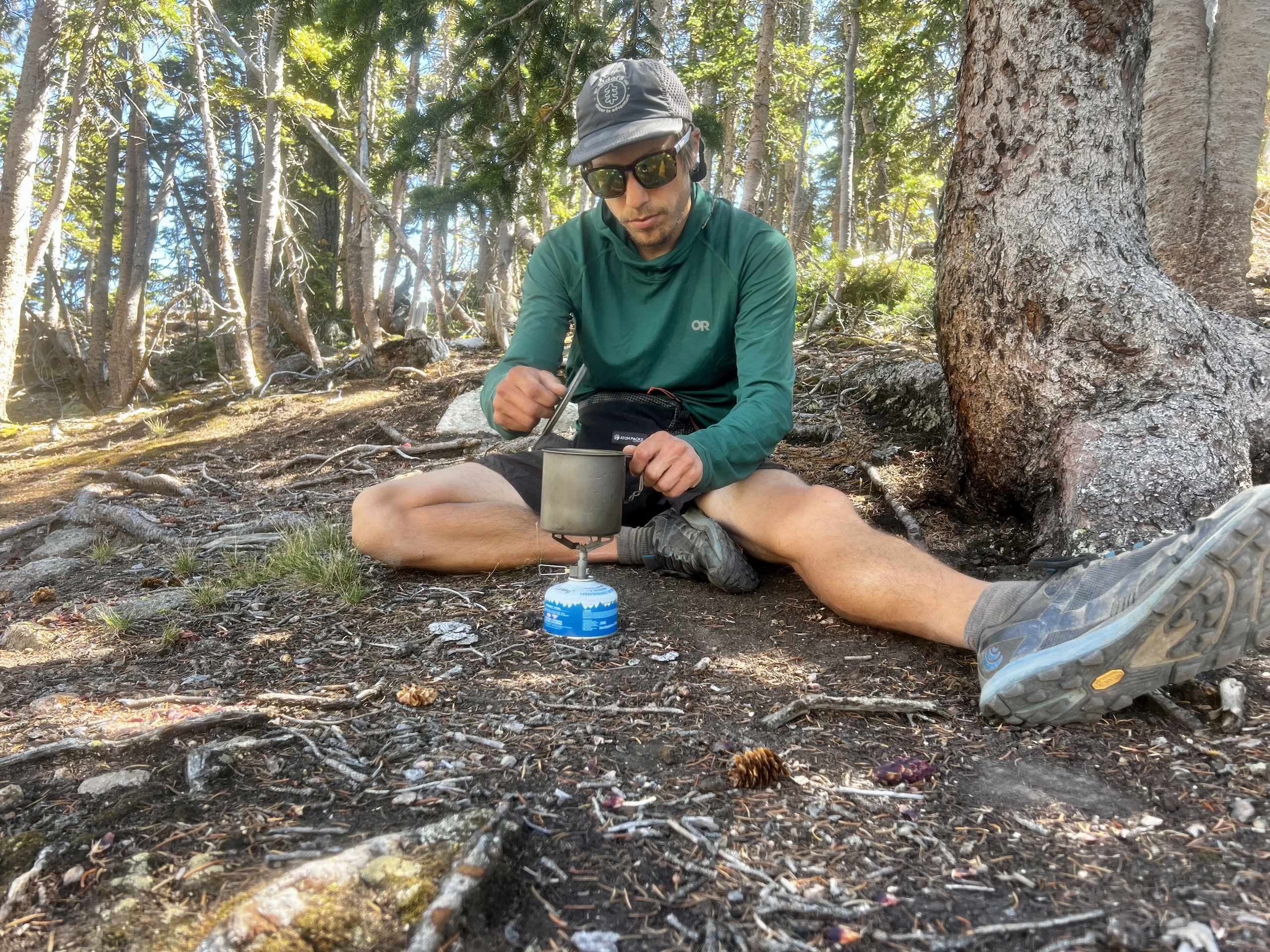 A hiker lifts the lid of his pot while cooking with the BRS 3000T in a wooded setting.