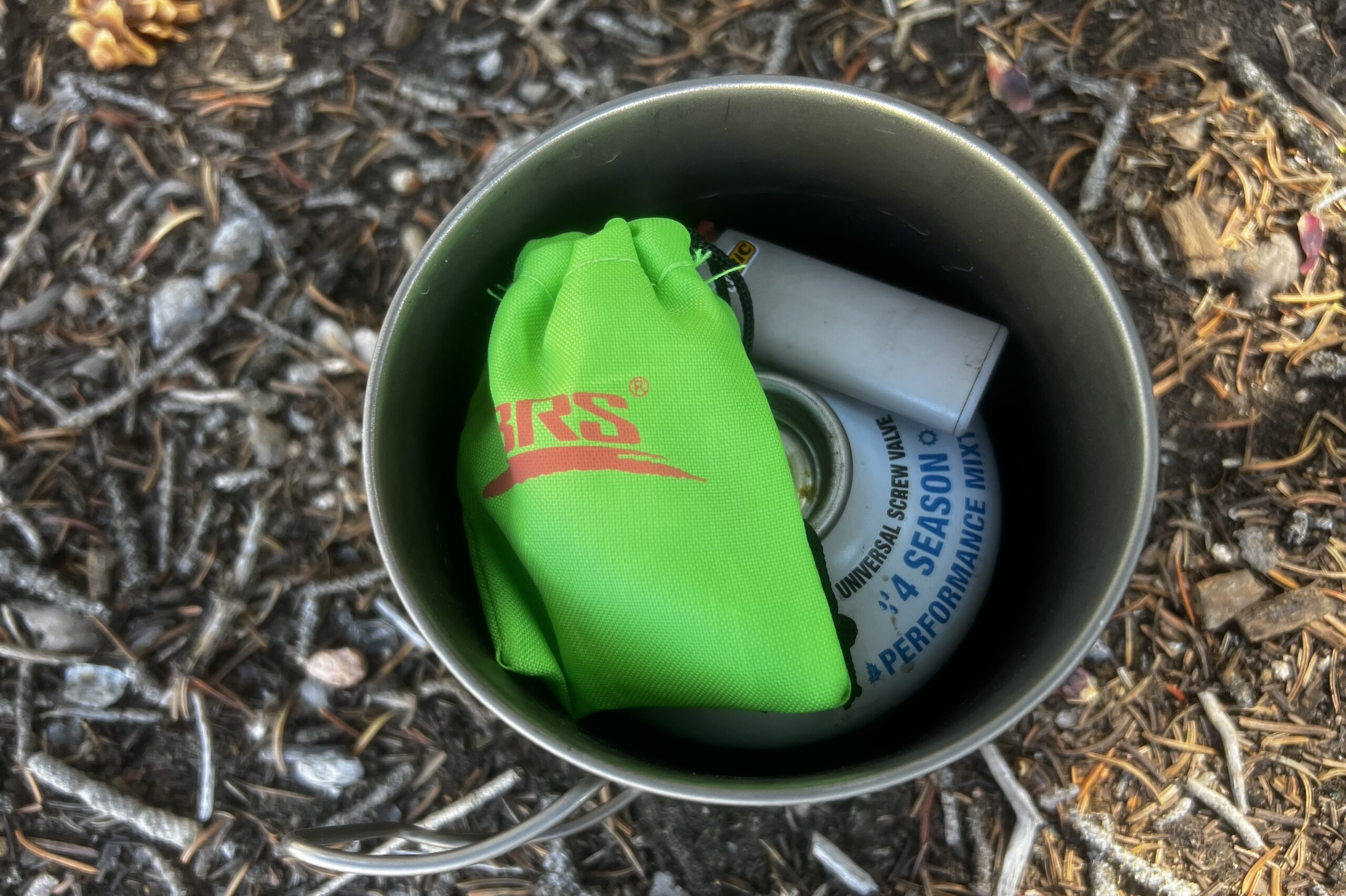 The BRS 3000T stove packed in a 550mL pot with stuff sack, an 8oz fuel canister and mini lighter. The whole kit sits on a pine needle forest floor.