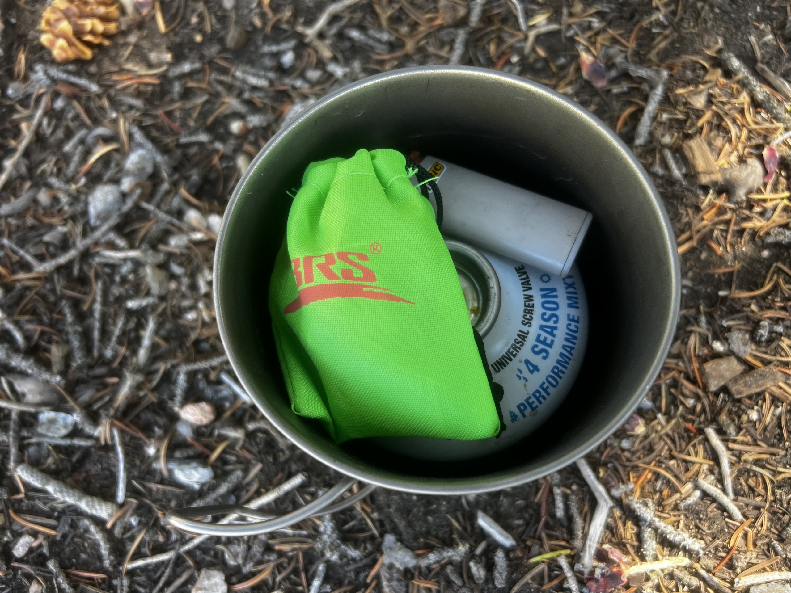 O fogão BRS 3000T embalado em uma panela de 550 mL com saco de material, um recipiente de combustível de 8 oz e mini isqueiro. O kit inteiro fica no chão de uma floresta de agulhas de pinheiro.