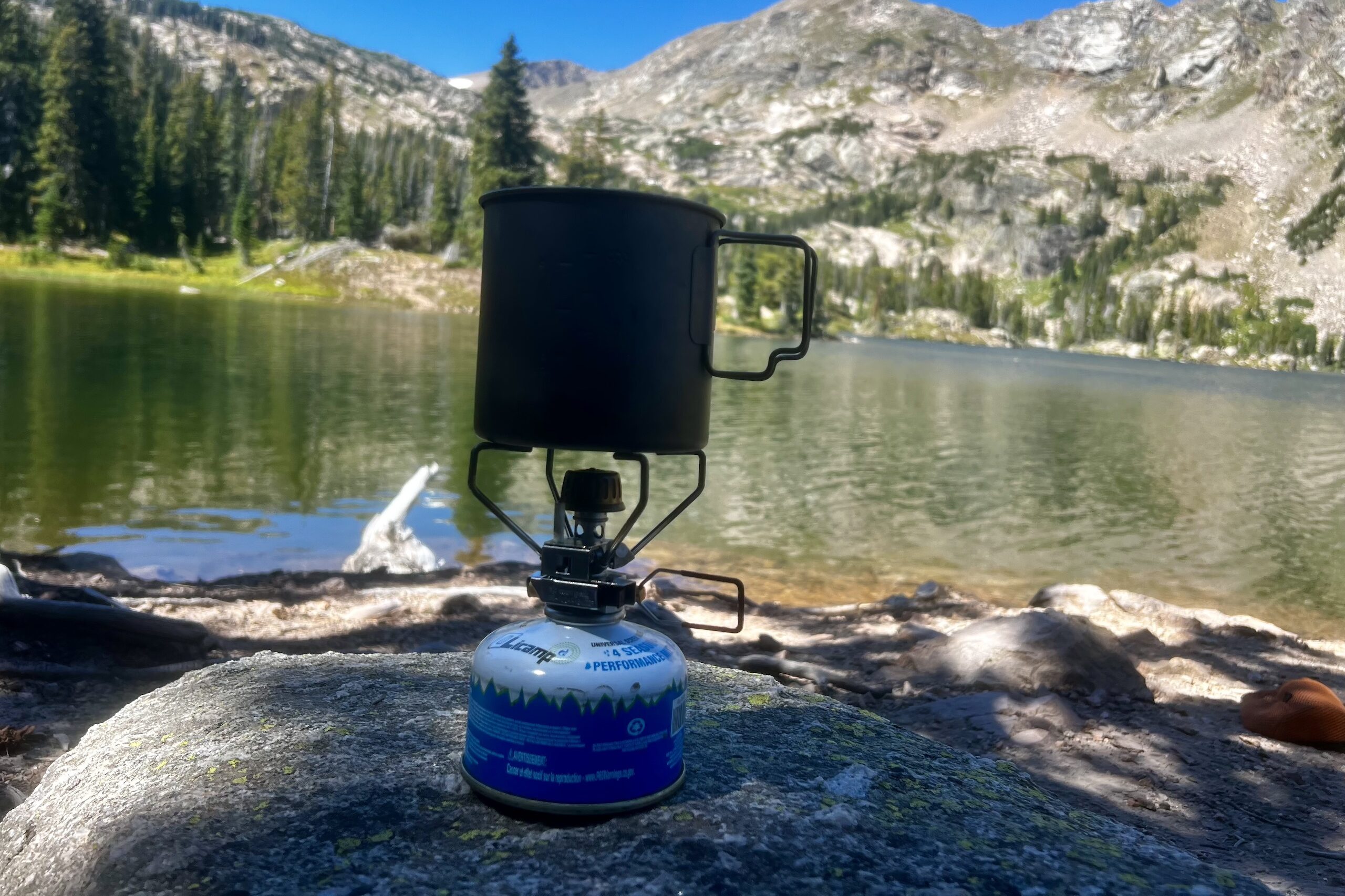 The Snow Peak Gigapower 2.0 sits on a rock, installed on a fuel canister, with a pot on top. The background is mountains and a lake.