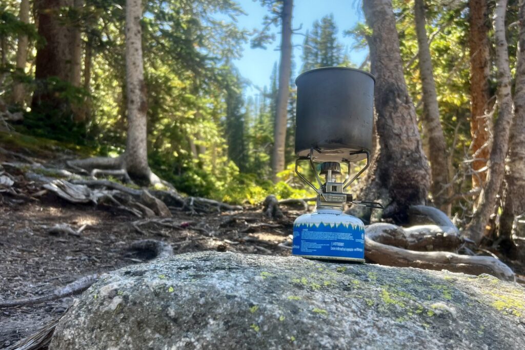 A low angle view of the Snow Peak Gigapower 2.0 sitting on a rock with trees in the background. The stove is installed with a small fuel canister, and a small cook pot is on the pot stand.