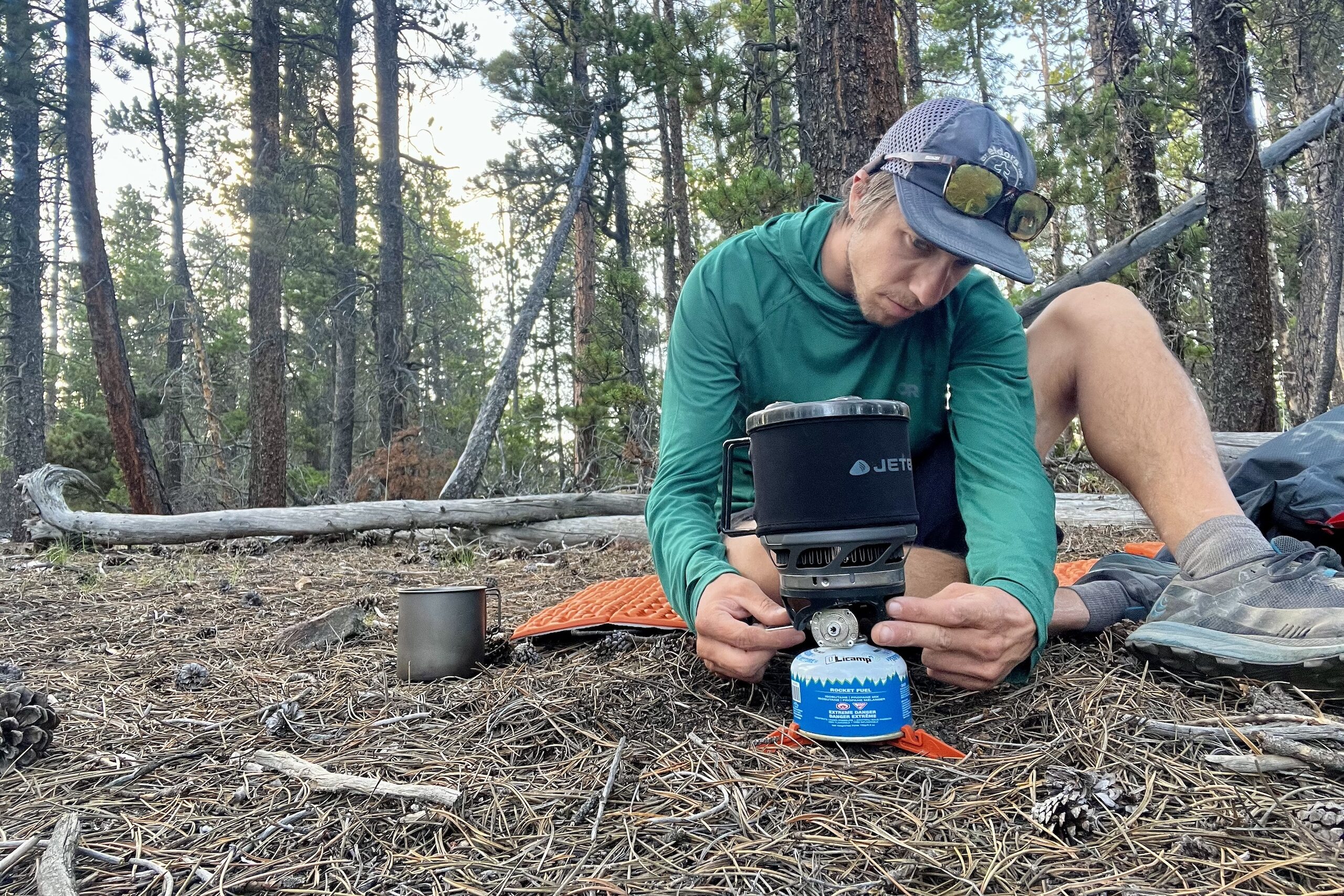 A hiker sits on the forest floor with his JetBoil MiniMo. He is turning the fuel valve with one hand while pushing the igniter switch with the other. Trees in the background.