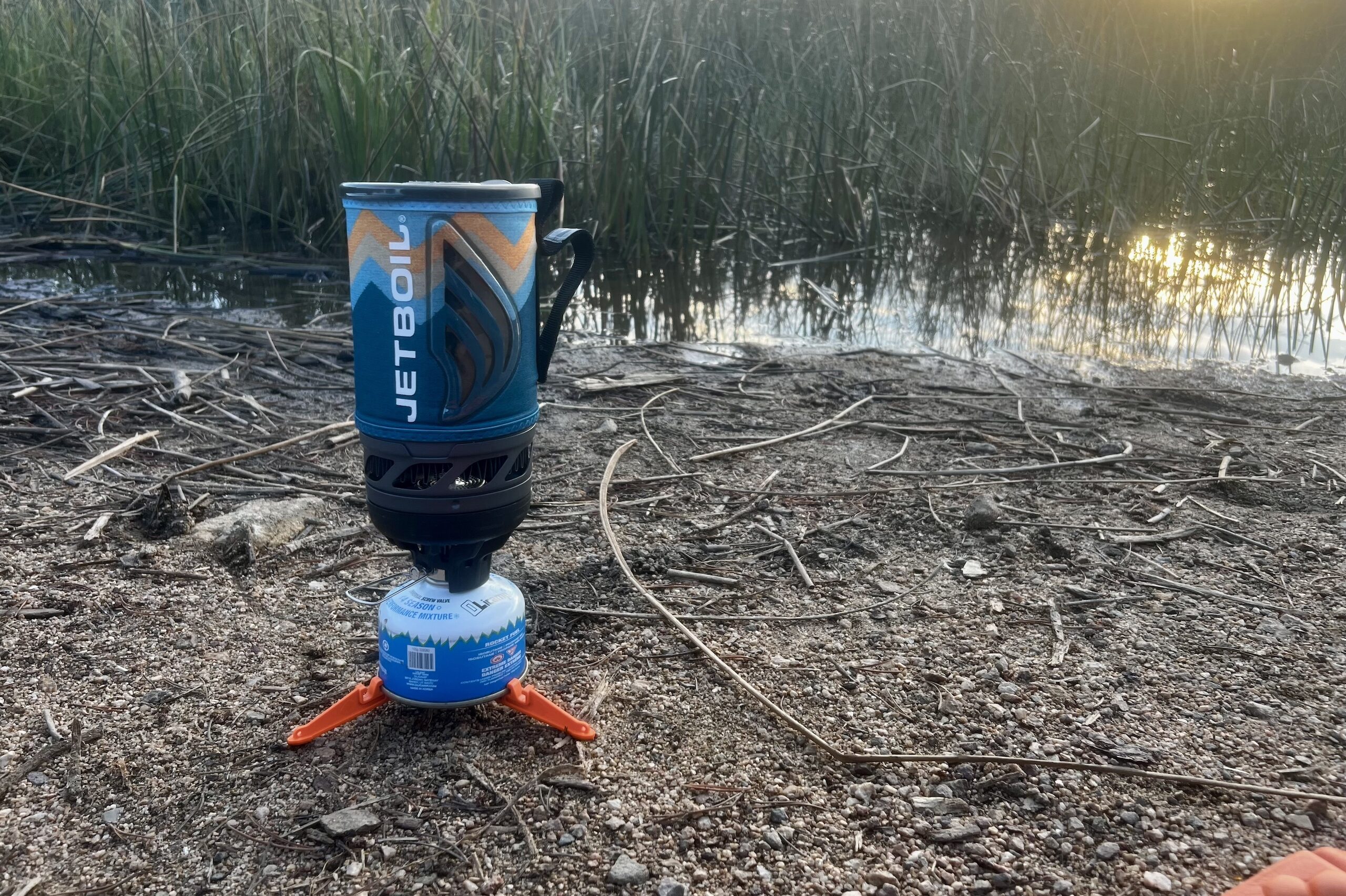 The Jetboil Flash sits on the ground, installed on a fuel canister and canister support in front of tall grass, water, and sunset.