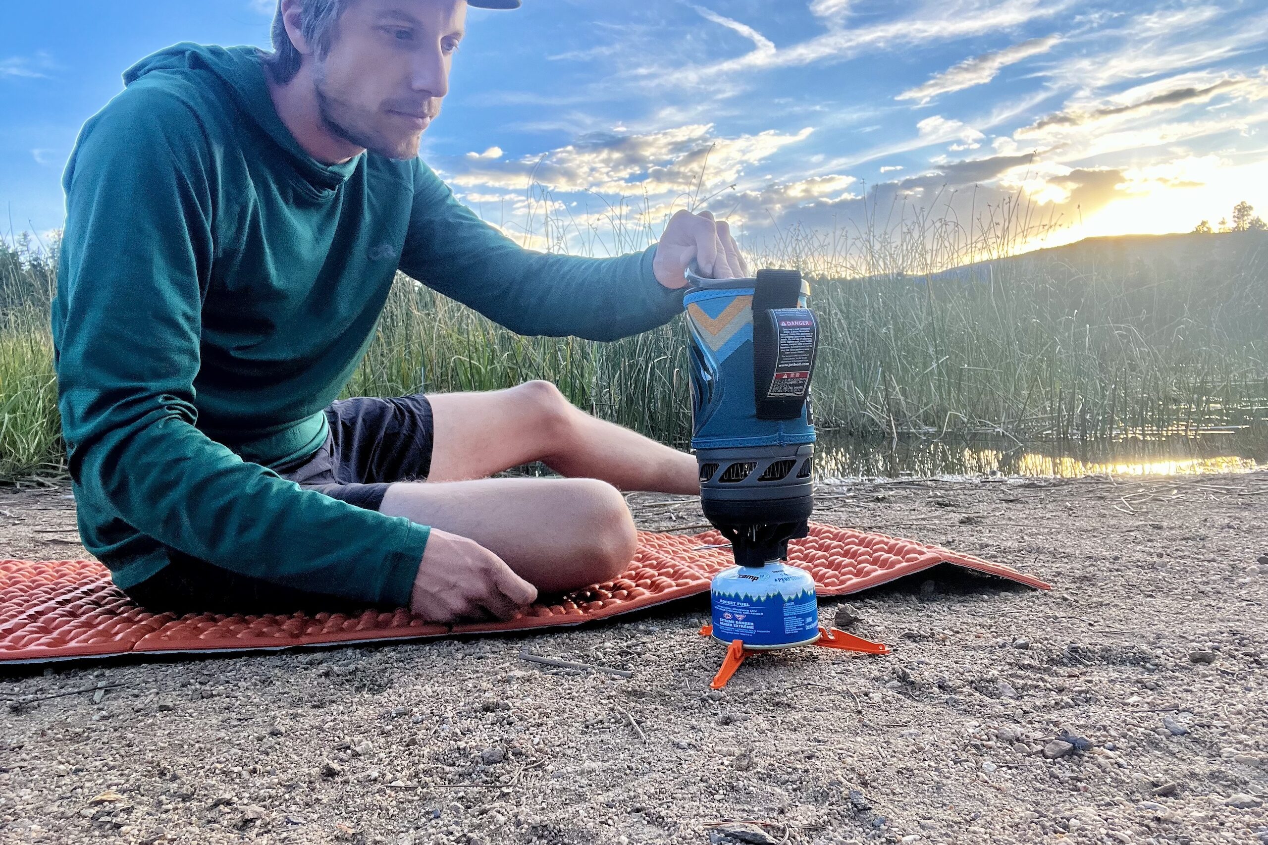 A hiker sits on a foam mat, cooking with the jetboil flash during sunset near tall grass. He is peeling the plastic lid off the jetboil which is sitting on the sand next to him.