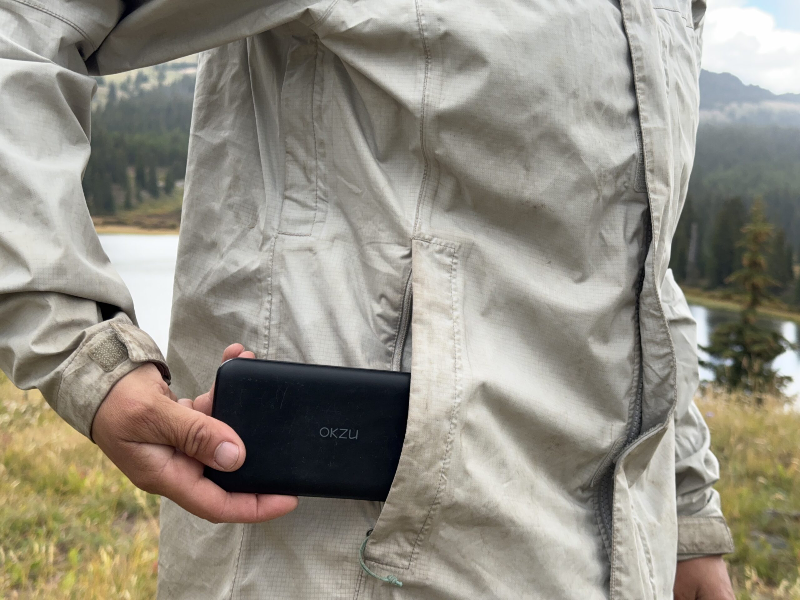 A hand placing the OKZU into a rain jacket pocket with mountains in the background.