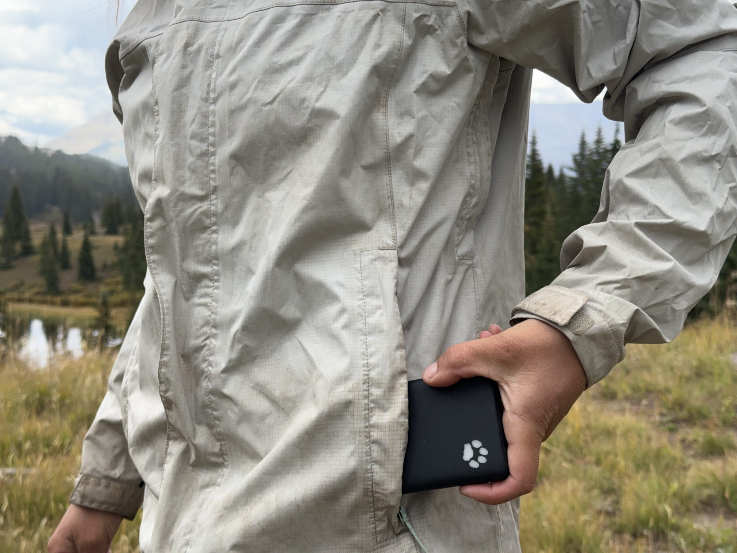 A hand placing the INIU Megapower 10000 into a rain jacket pocket with trees and mountains in the background.