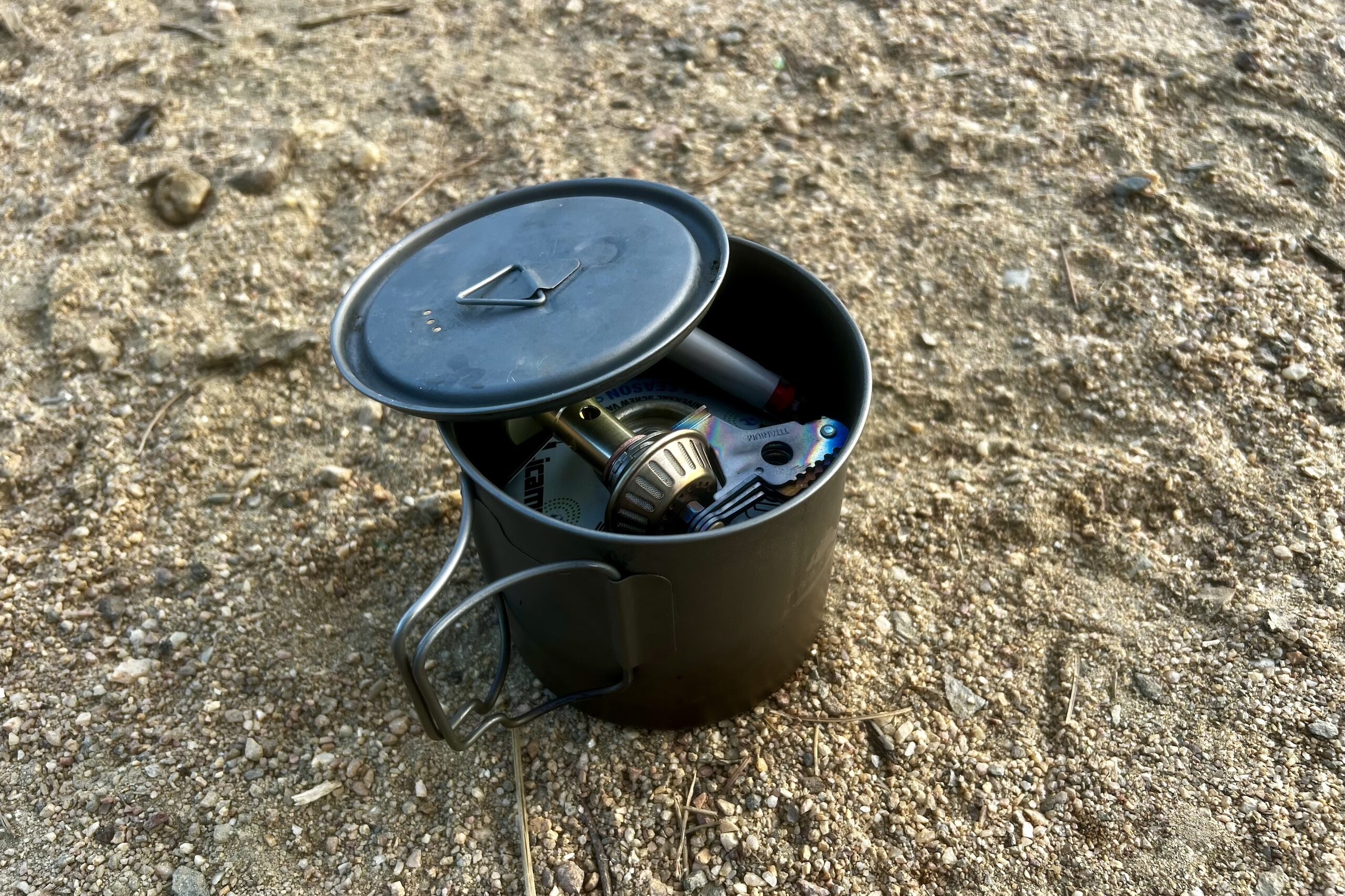 The Snow Peak LiteMax packed into a pot with fuel and lighter and the lid hanging off the side. It sits on sandy soil.