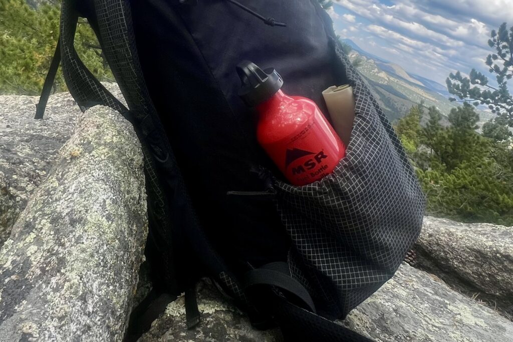 An MSR fuel bottle packed into the side water bottle pockets of a backpack. Rocks and mountains are in the background.