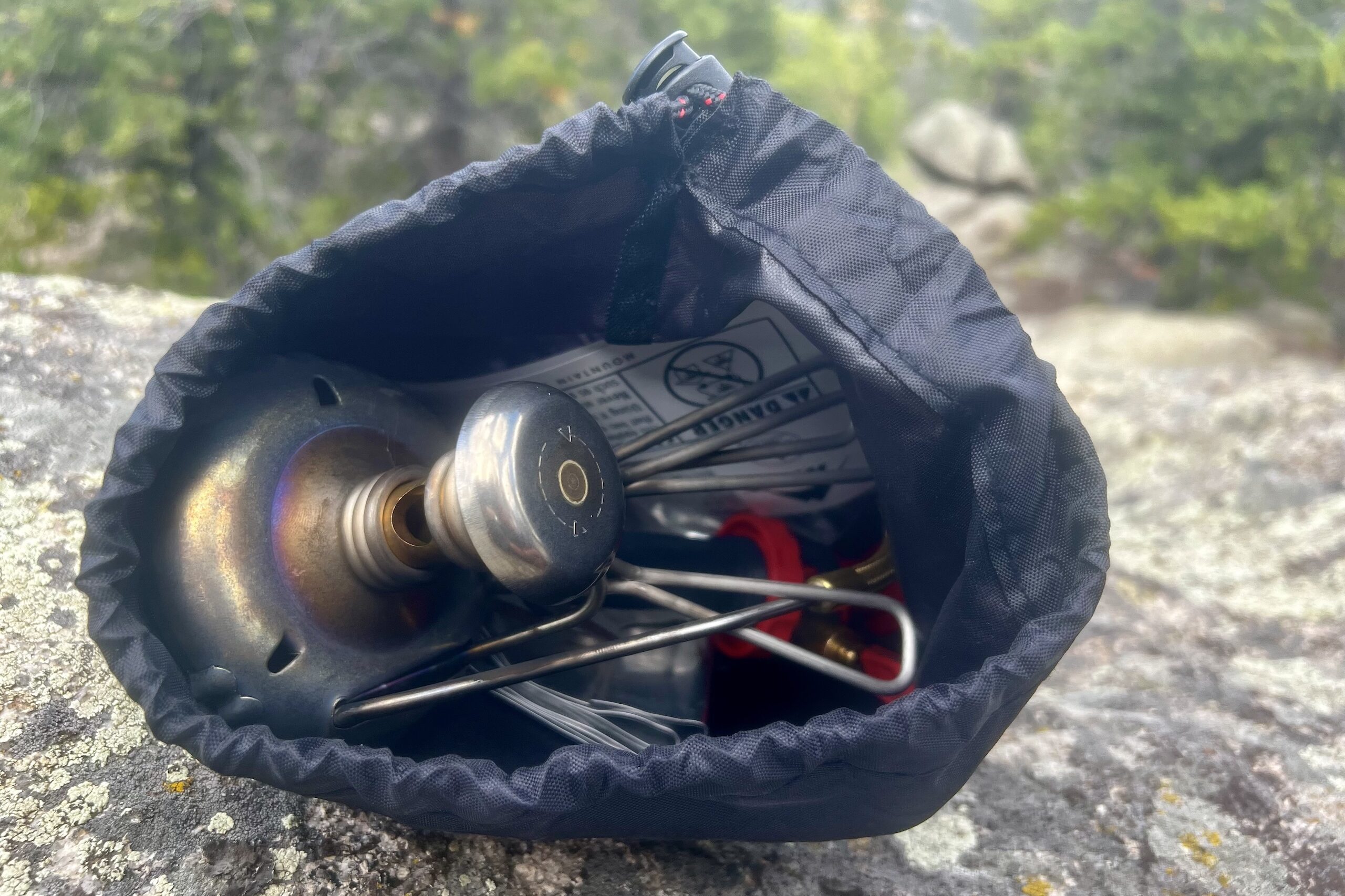 A view of the MSR Whisperlite and all of its components packed into its stuff sack. Trees in the background.