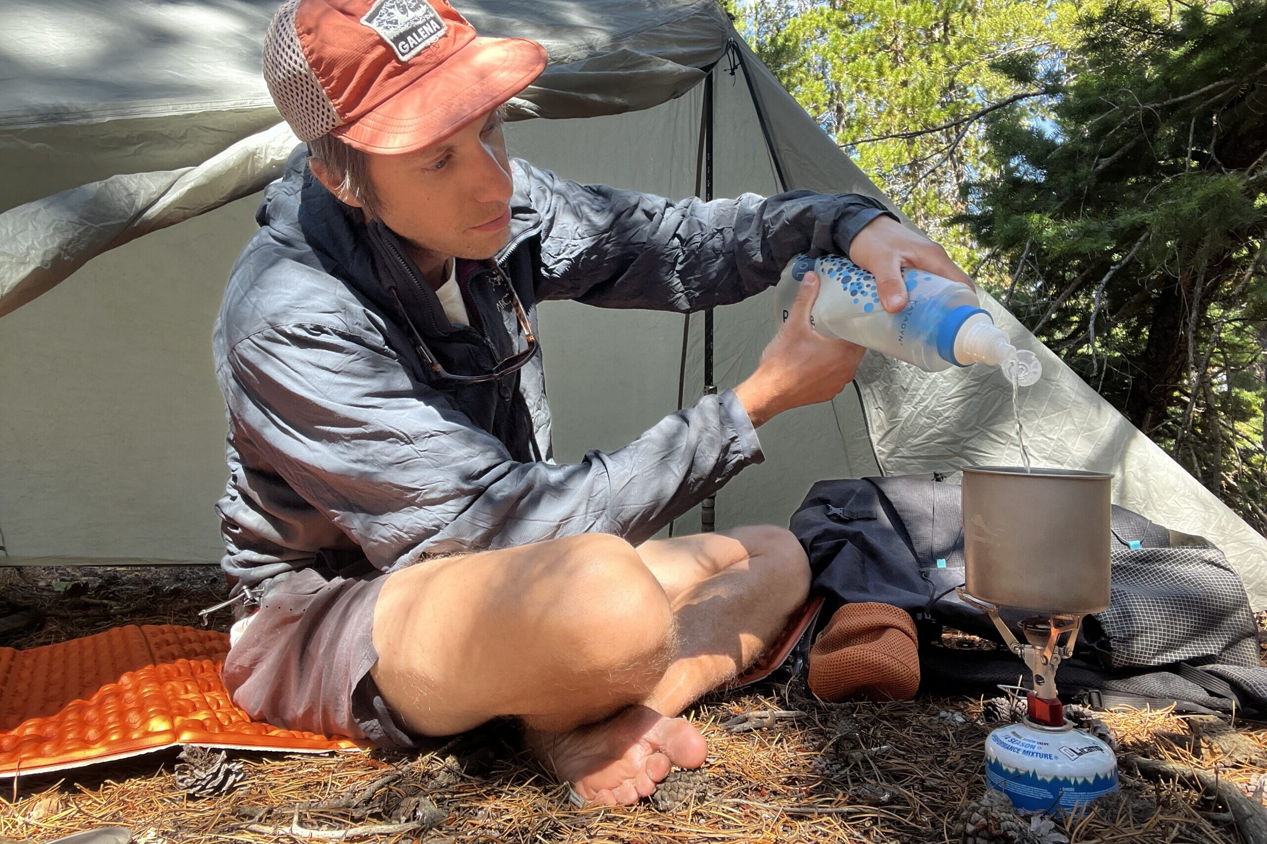 A hiker filters water into a pot that is sitting on the MSR PocketRocket 2. Tent, backpack, and trees are in the background.