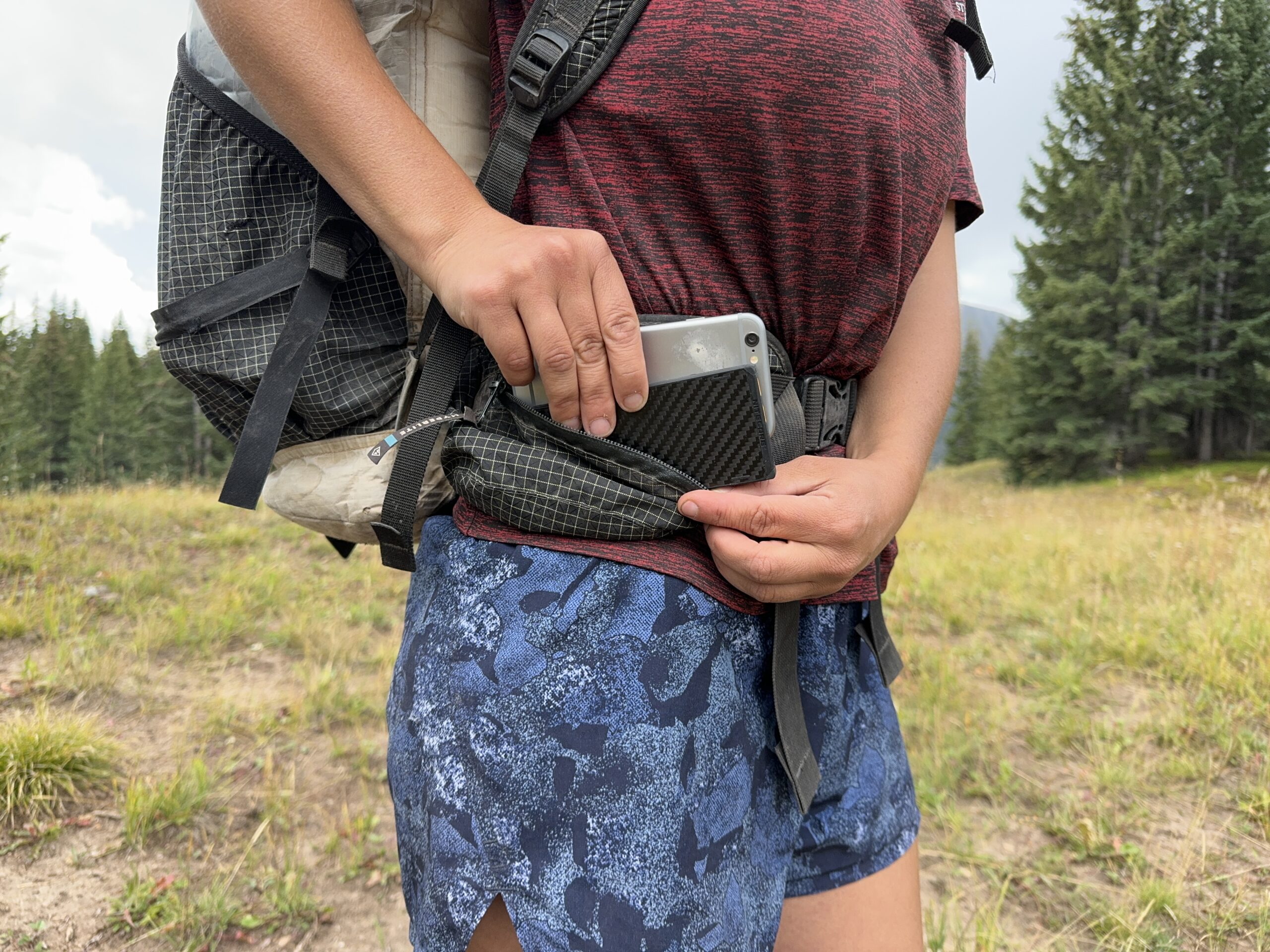 Close up of a backpack side pocket with one hand placing the Nitecore and a cell phone into the pocket and the other holding the zipper. There are trees in the background.