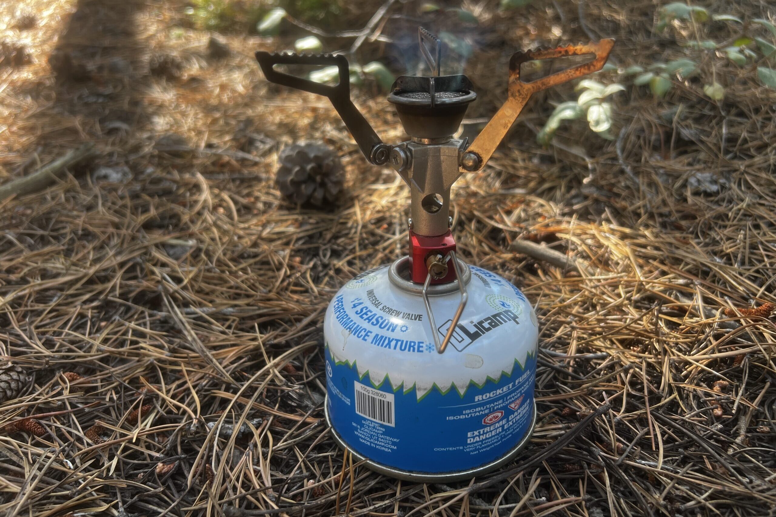 The MSR PocketRocket 2 sits on the ground with installed fuel canister and the flame on. Plants and pine needles are in the background.