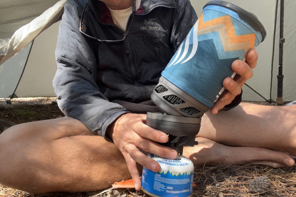 A hiker at camp, sitting in front of a tent, screwing the JetBoil Flash pot onto its burner. One hand stabilizes the burner and fuel while the other hand tilts the bottom of the stove into the burner to screw it in place.