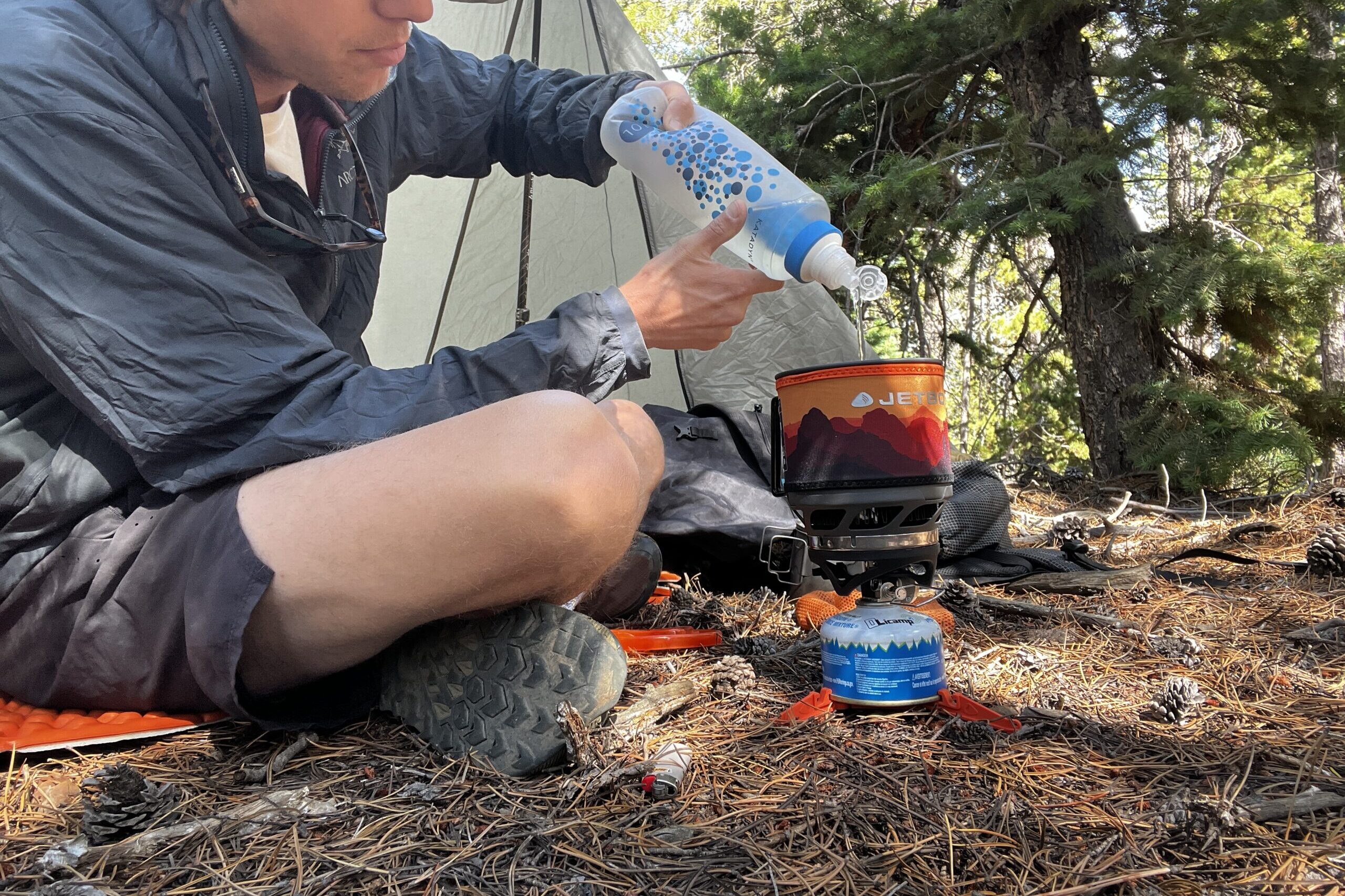 A hiker filters water into his MiniMo pot. The pot is installed with its burner, fuel canister, and canister support. The background has trees and backpacking gear.