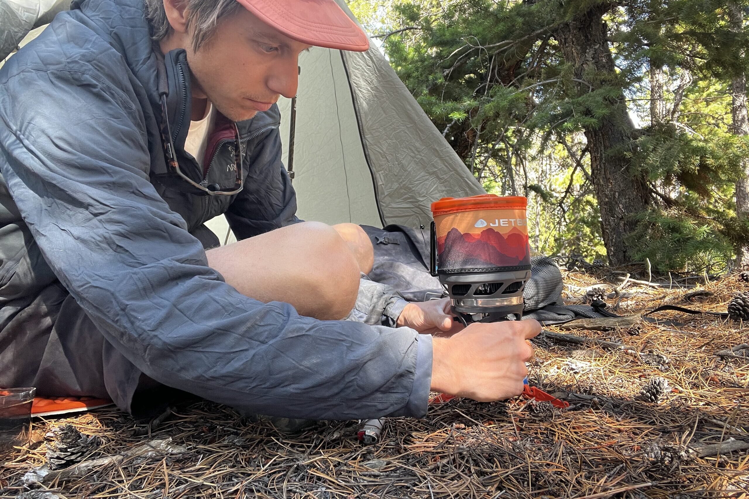 A hiker gets ready to light his MiniMo. He is leaning over with both hands on the base of the stove. Trees and backpacking gear are in the background.