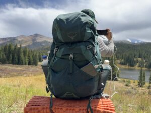 A hiker has their back to the camera while taking a photo. The phone is hooked up to the UGREEN charger in the back pocket of the pack. There are trees and a mountain lake in the background.