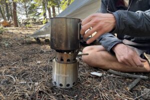 A hiker sets a pot on the Solo Stove Lite. A tent and trees are in the background.