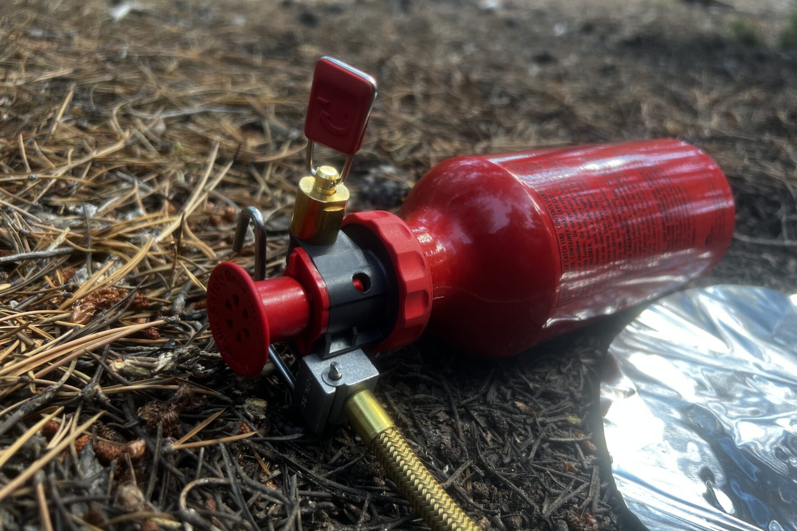 A closeup of the MSR Whisperlite fuel pump installed on a fuel bottle, sitting on a forest floor. The fuel line a heat reflector are partially in the photo.