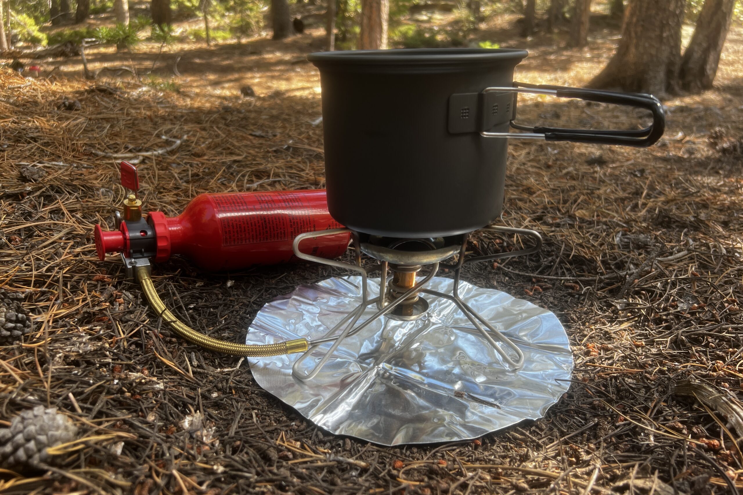 The Whisperlite stove with pot, heat reflector, and fuel bottle, all on a forest floor with trees in the background.