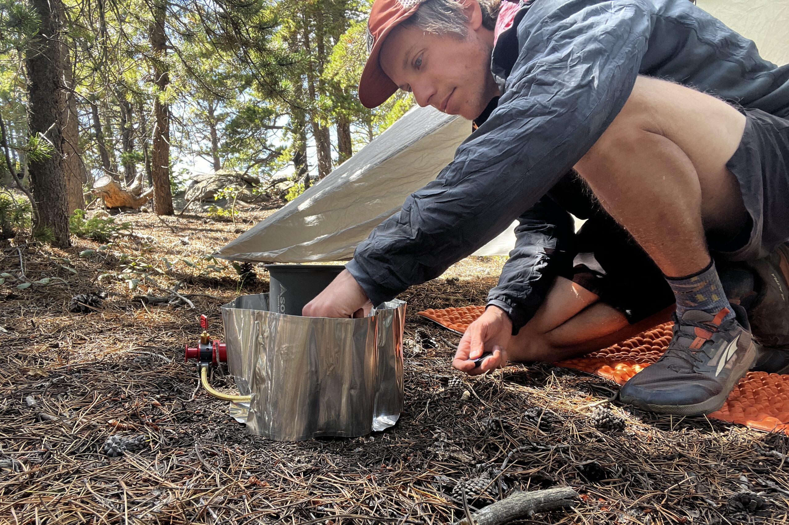 The Whisperlite sits on the ground with a pot on top and its windshield wrapped around. A hiker reaches down to light the priming cup of the stove. A tent and trees are in the background.