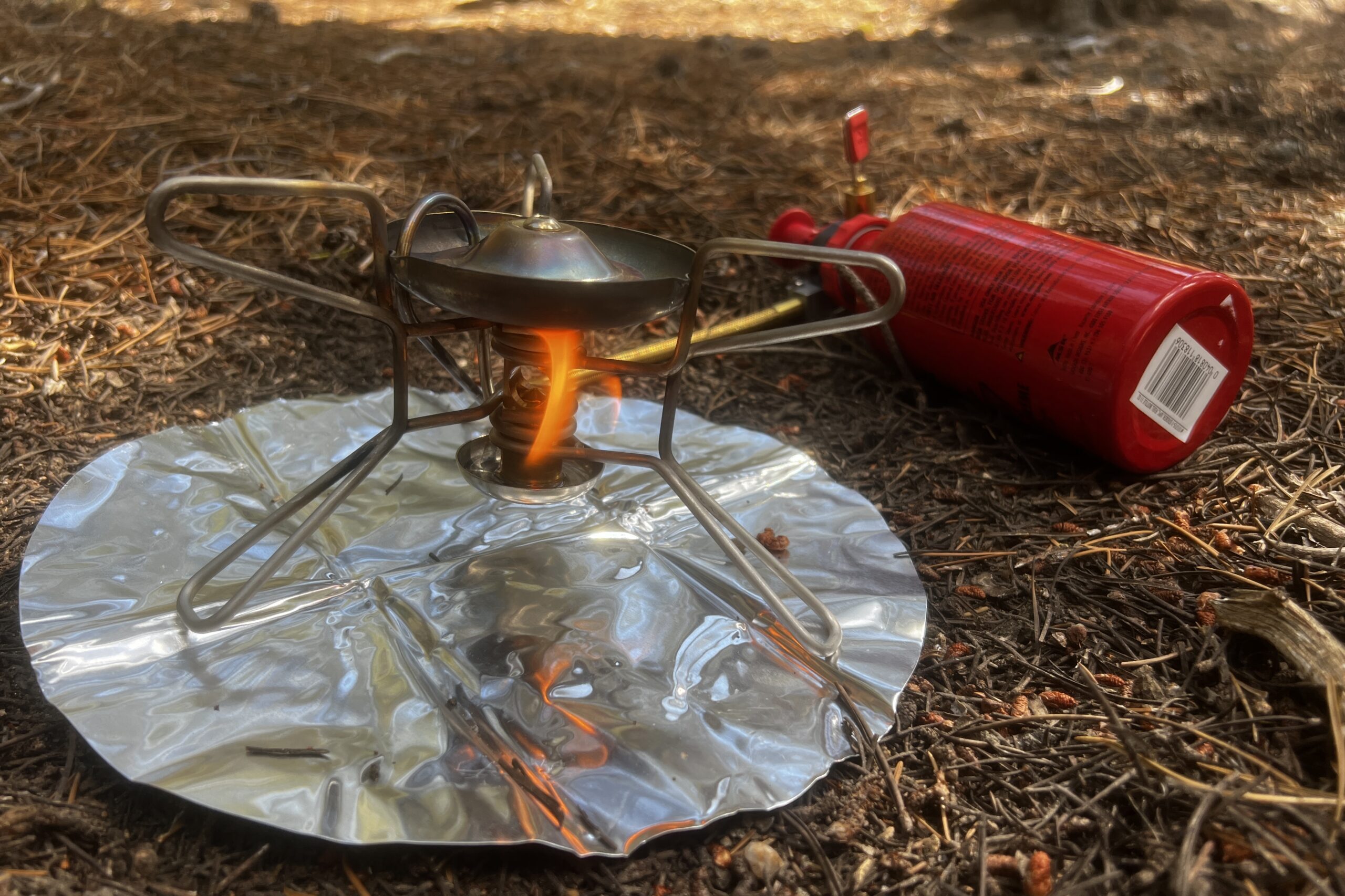 The Whisperlite sits on it's heat reflector with a fuel bottle attached. A flame is present, priming the main burner.