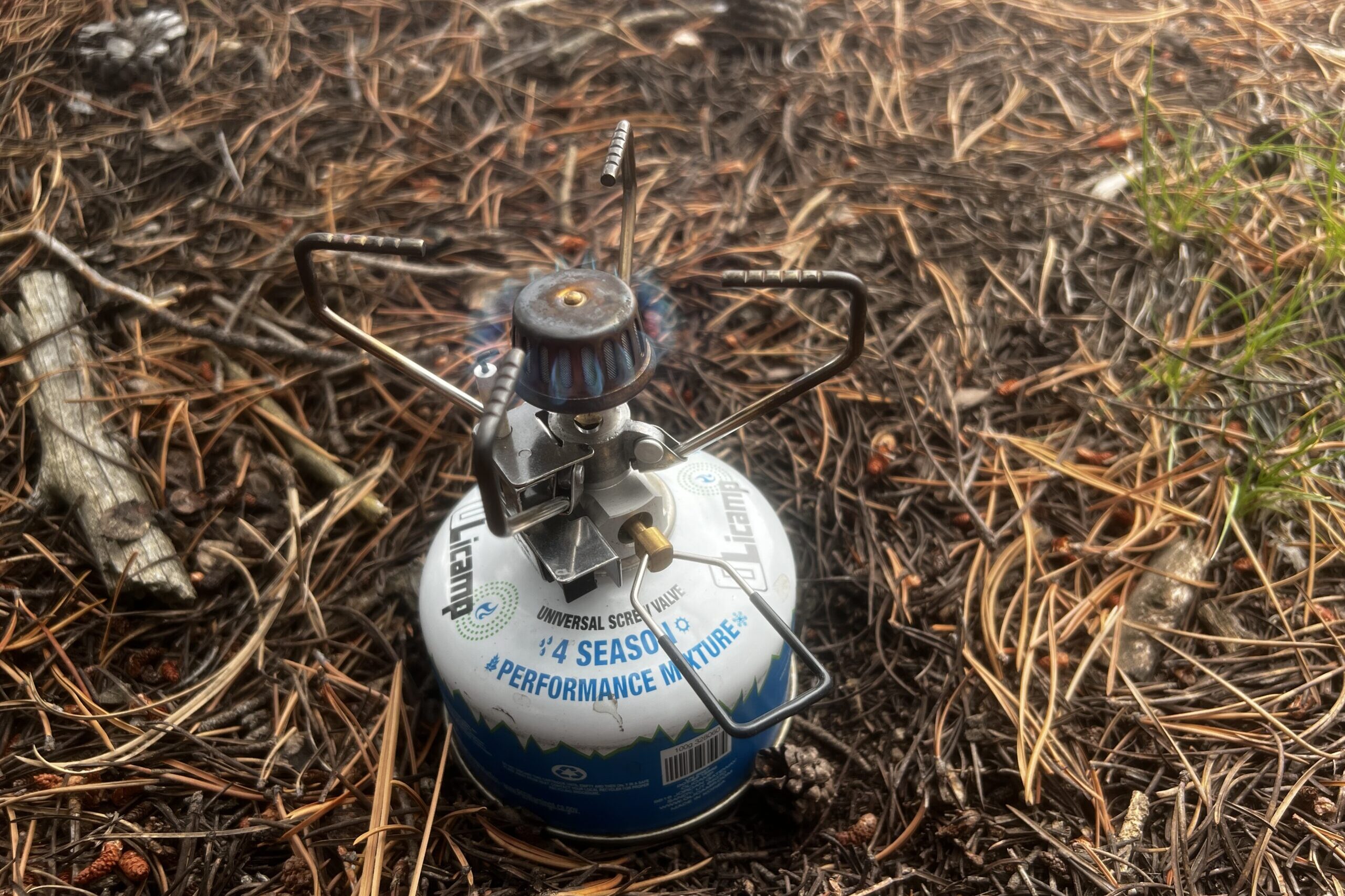 The Gigapower stove sits on a bed of pine needles. It is screwed onto a fuel canister and the flame is on.