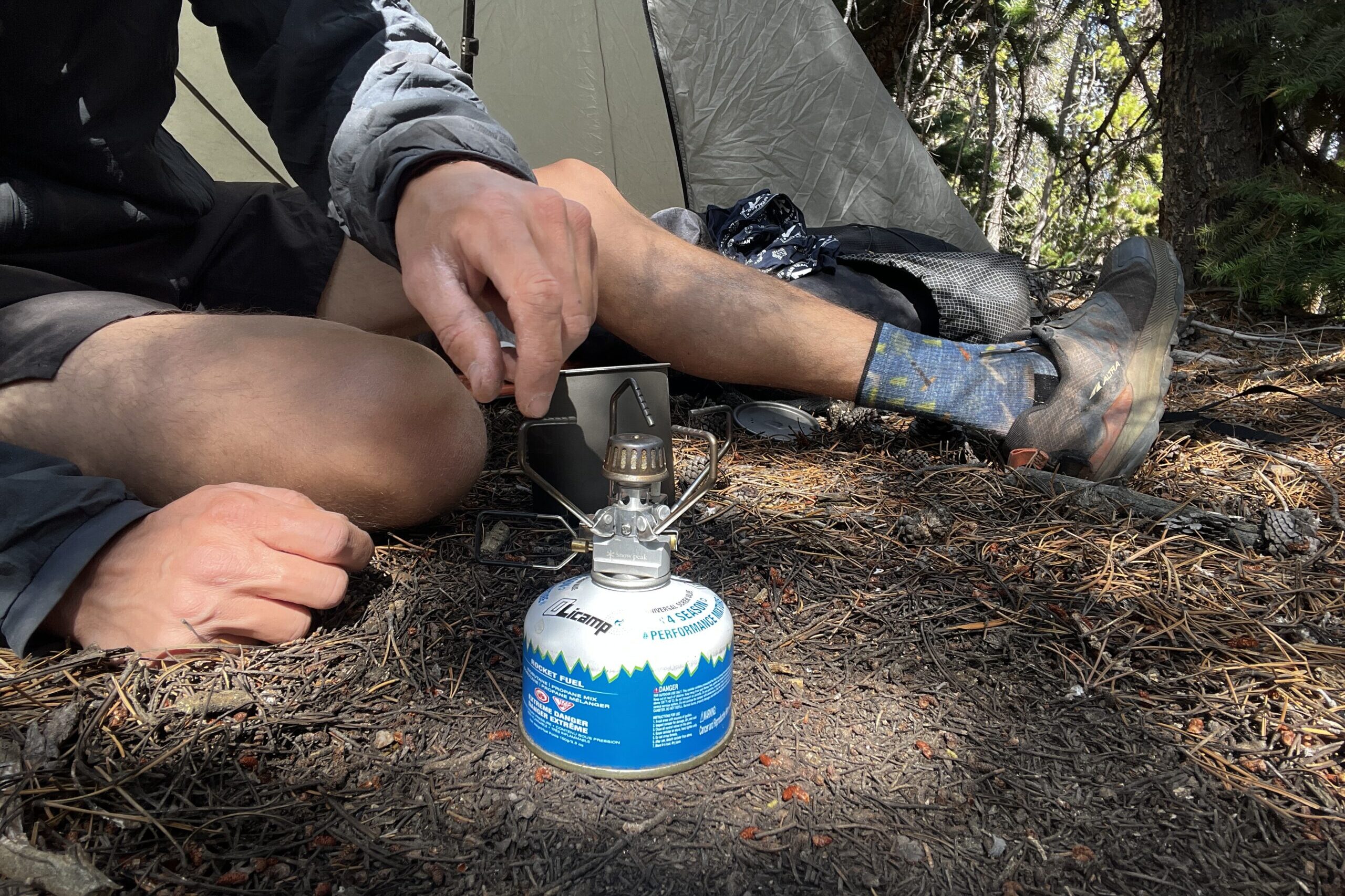 A hiker carefully unfolds the arms of the Gigapower 2.0. The stove is installed on a fuel canister and sits on the ground. The background includes a tent, camping gear, and trees.